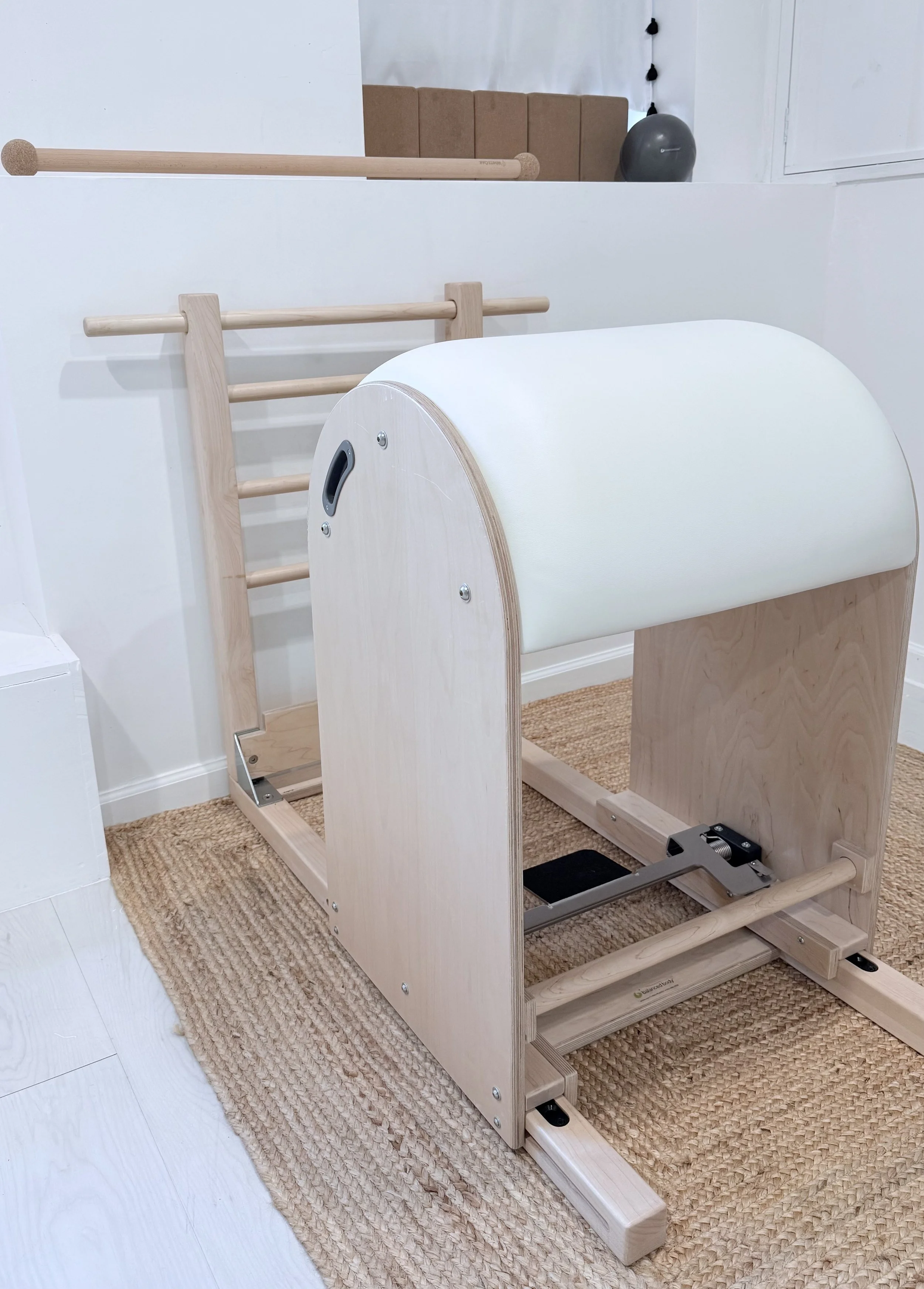 A wooden treadmill with a padded roller at the top, set on a woven rug in a room with white walls and various fitness equipment in the background.