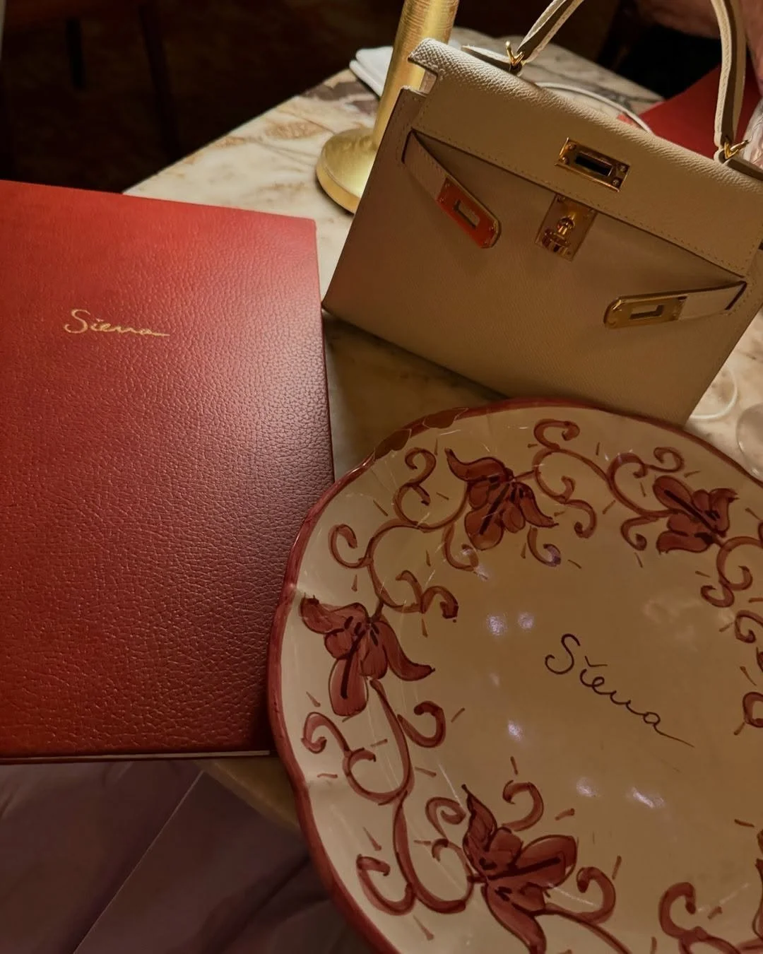 Une assiette décorative, un menu rouge, et un sac beige sur une table en marbre, avec un candélabre en arrière-plan.