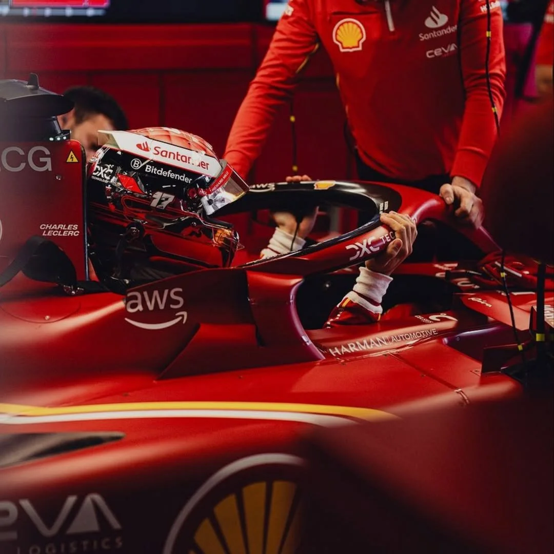 Un pilote de Formule 1 en tenue de course dans une voiture rouge, en train de se préparer dans un garage, avec un casque blanc et le numéro 16 visible.