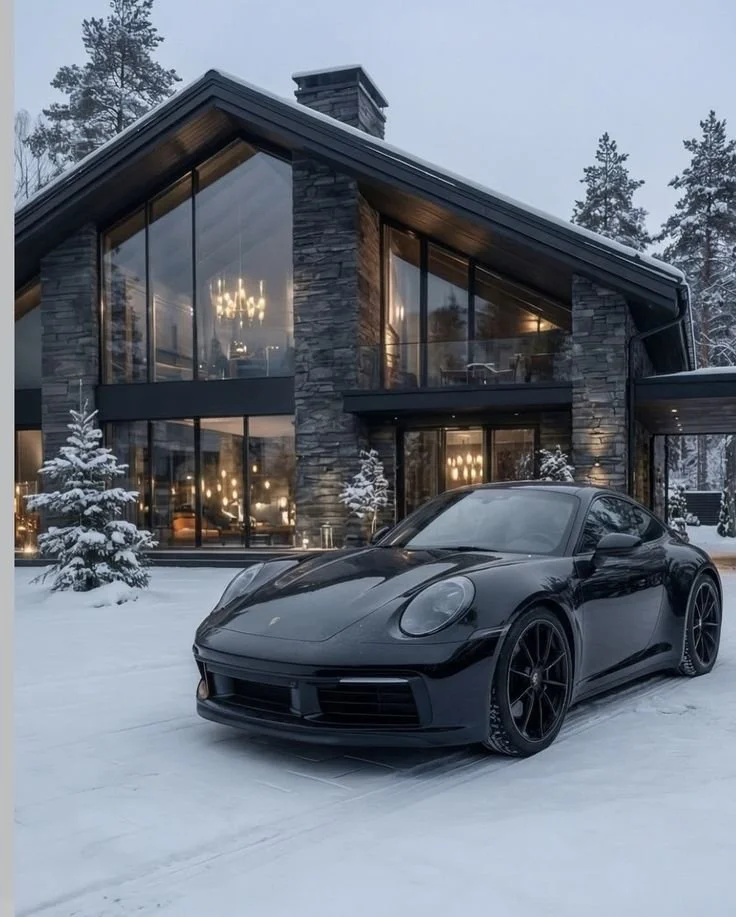 Voiture de sport noire devant une maison en pierre avec de grandes fenêtres, dans un paysage enneigé.