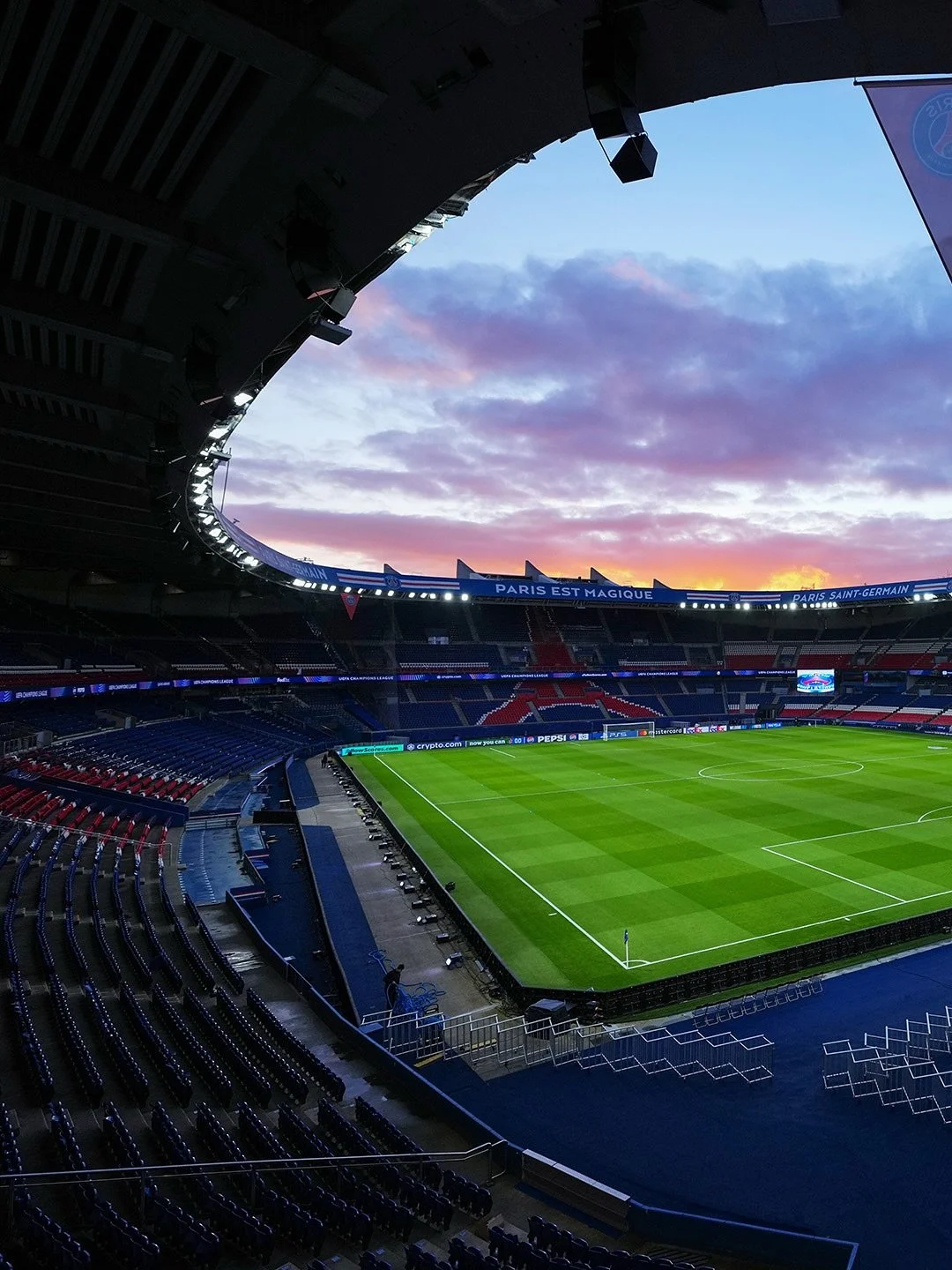 Une vue d'un stade de football vide avec un ciel coloré lors du coucher du soleil, des sièges rouges et bleus, et un terrain bien entretenu.