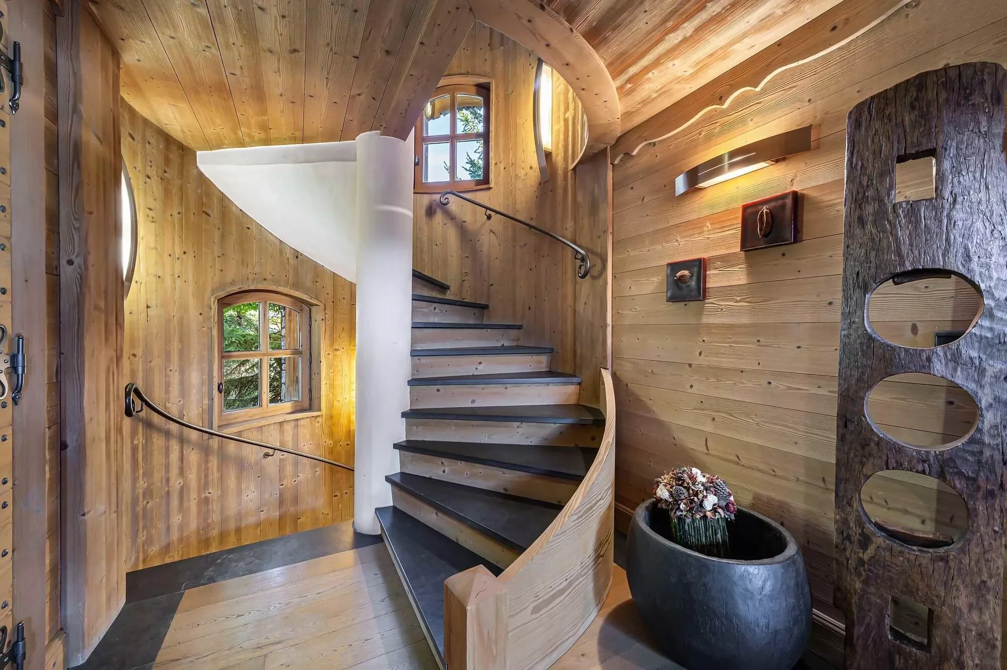 Escalier hélicoïdal en bois et métal dans un intérieur de chalet en bois, avec fenêtres et grand pot décoratif.