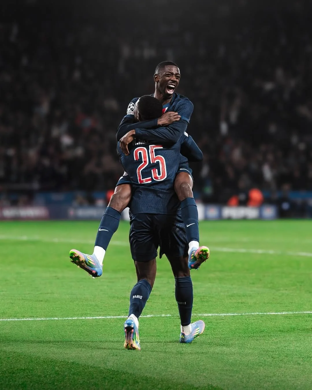 Deux joueurs de football en uniforme célébrant une victoire sur le terrain, un en sautant sur l'autre, sous les projecteurs.