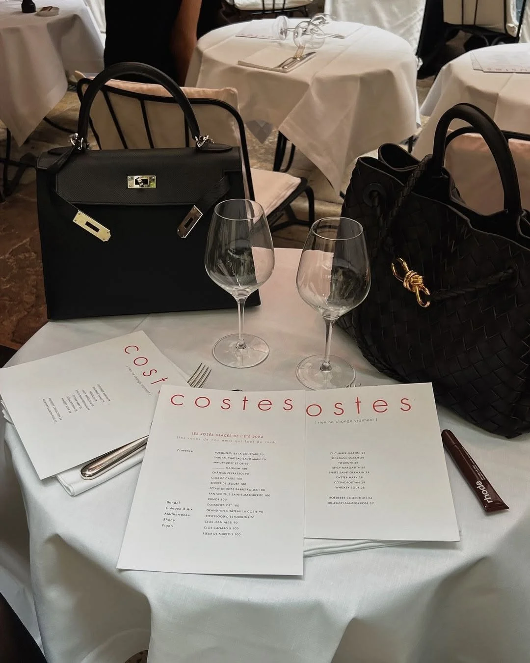 Une table de restaurant élégante avec deux sacs à main noirs, deux verres à vin, des menus et des couverts, dans un cadre chic avec des chaises en métal et des nappes blanches.