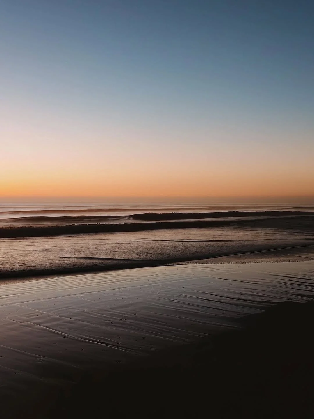 Plage déserte au coucher du soleil, avec des vagues douces et du sable humide réfléchissant la lumière.