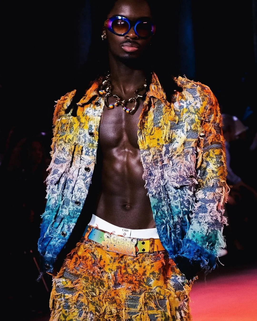 Un homme portant des vêtements colorés et déchirés, avec des accessoires de mode tels que des lunettes de soleil et des bijoux, défilant sur un podium.