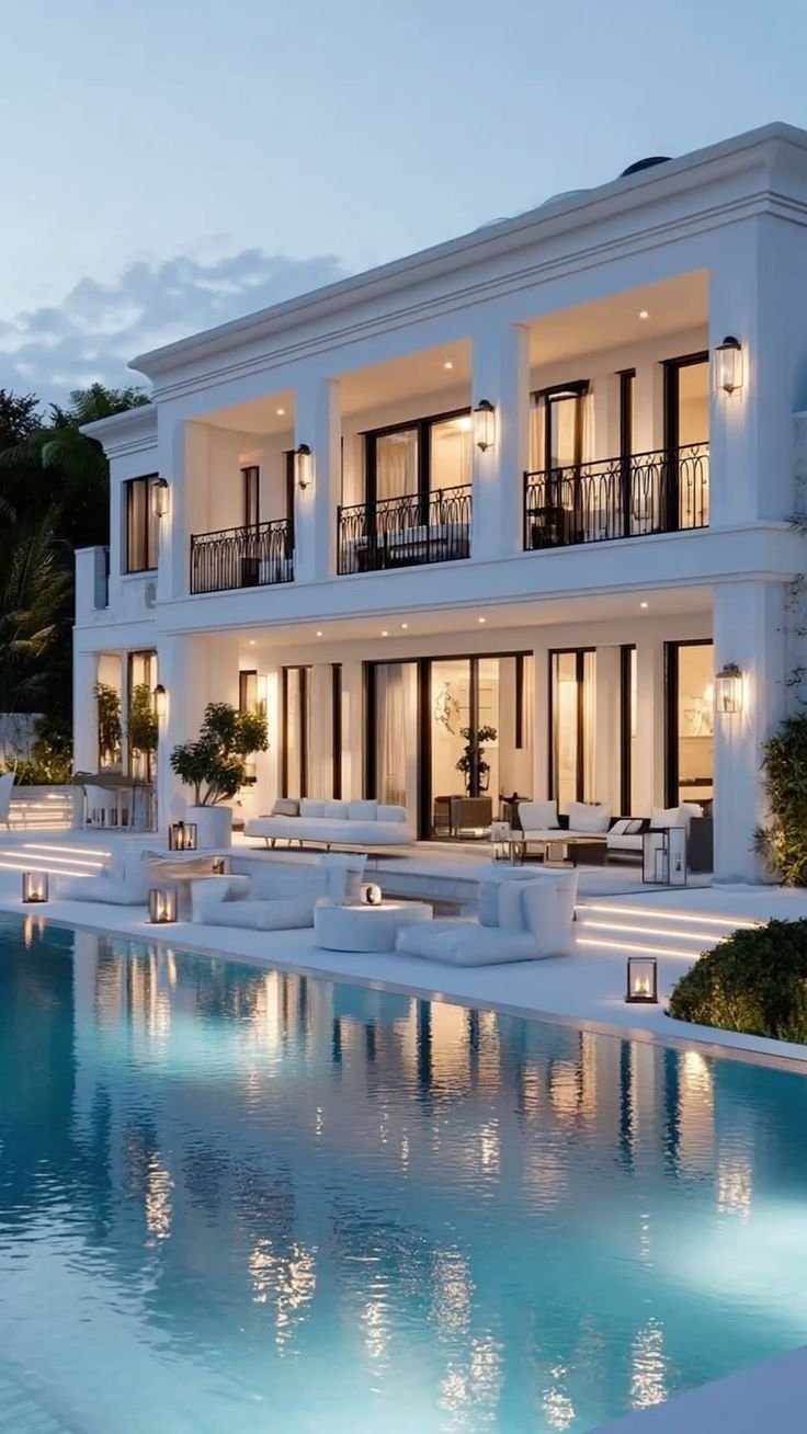Une magnifique maison moderne blanche avec deux étages, plusieurs balcons avec des balustrades en métal, des lumières extérieures, une terrasse avec des meubles en blanc, une piscine claire et des lanternes éclairant la scène, le tout dans un cadre é