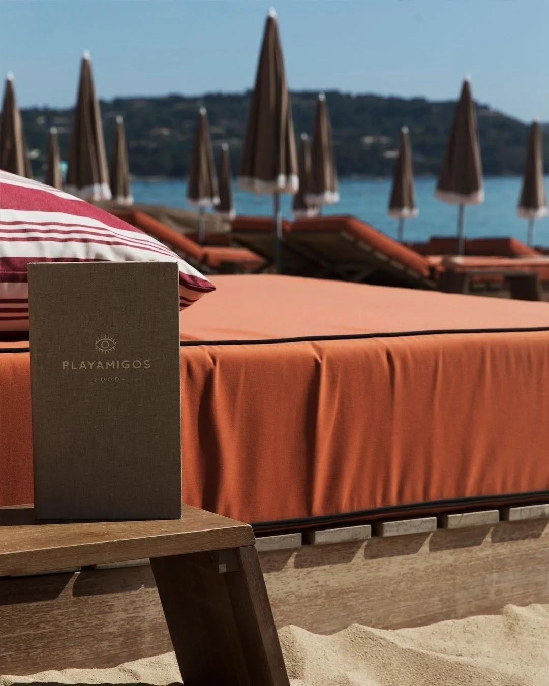 Une plage avec des lits de soleil et des parasols en tissu marron, un menu de restaurant sur une table en bois, la mer en arrière-plan, et une colline au loin.