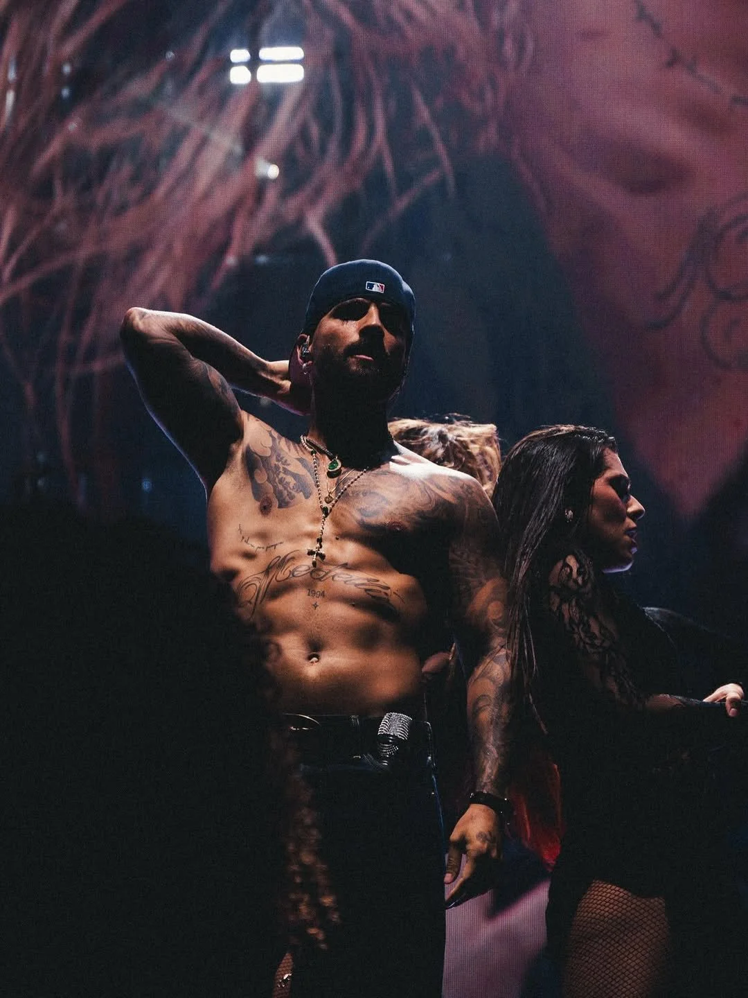 Un homme tatoué, torse nu, portant une casquette, se tient sur scène avec des personnes en arrière-plan dans un environnement de concert.