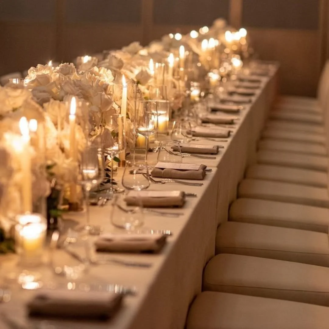 Table de réception élégamment dressée avec fleurs blanches, bougies allumées, verrerie et chaises beiges.