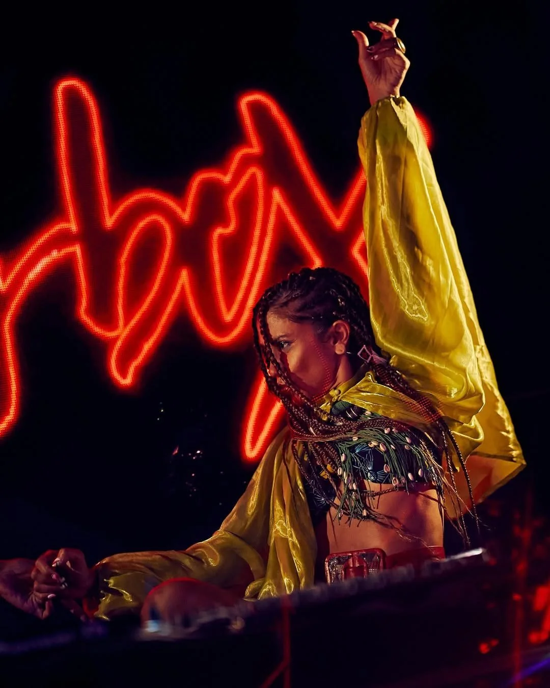 Une femme aux cheveux en tresses, portant une veste jaune en satin, fait une pose de danse ou de mannequin devant un panneau de néon rouge en forme de graphique ou d'onde acoustique.