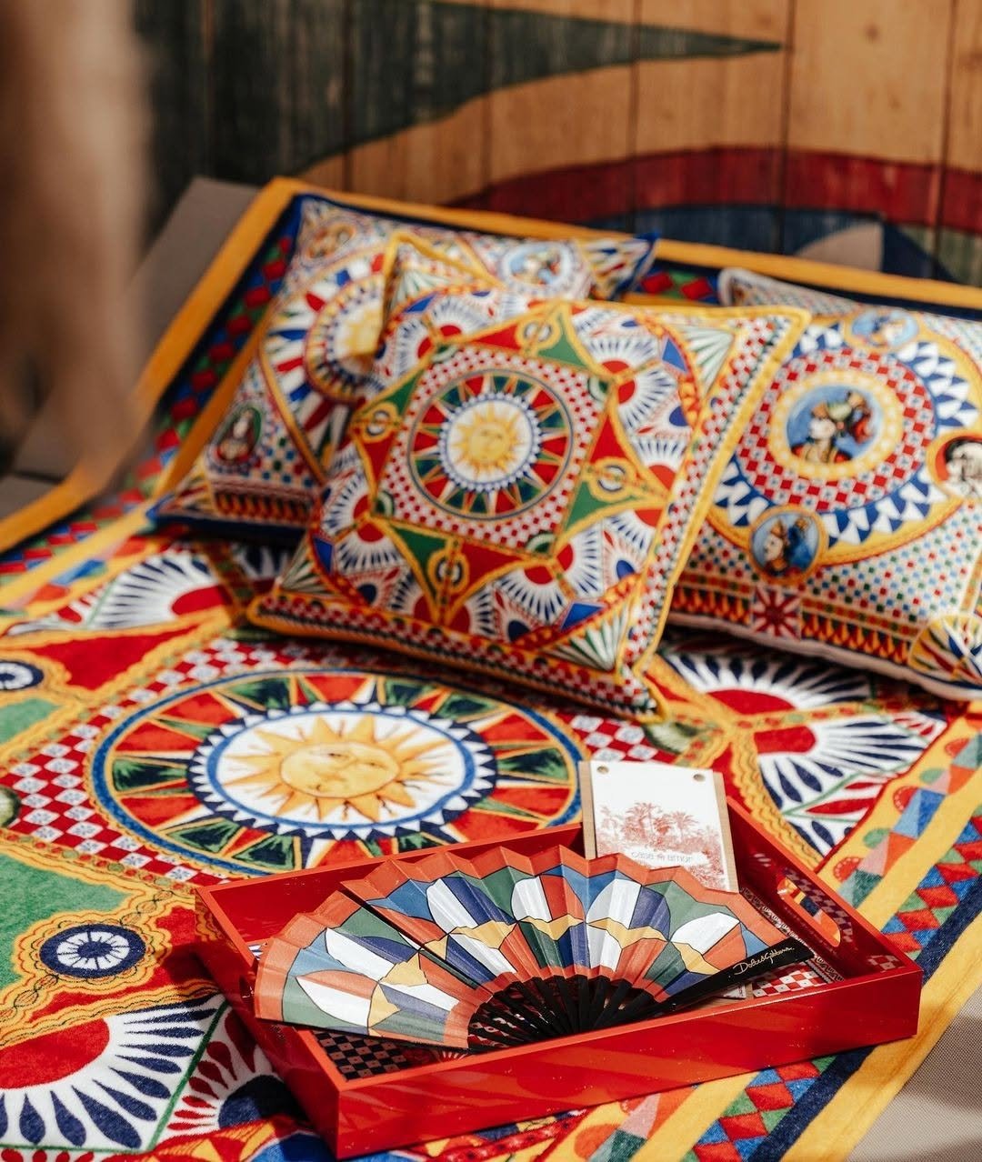 Coussins décoratifs colorés avec des motifs géométriques et des images de personnages, placés sur une surface recouverte d'un tissu avec un motif similaire, en arrière-plan il y a un mur en bois