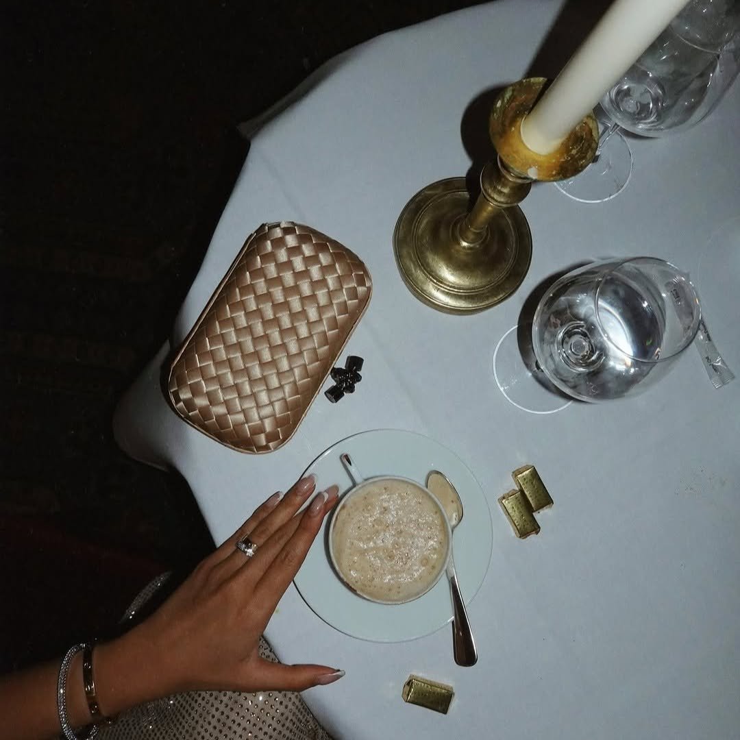 Une table ronde avec une bougie, deux verres d'eau, une main avec une bague et un thé ou café dans une tasse, un porte-monnaie en cuir synthétique et des petits cadeaux en or.