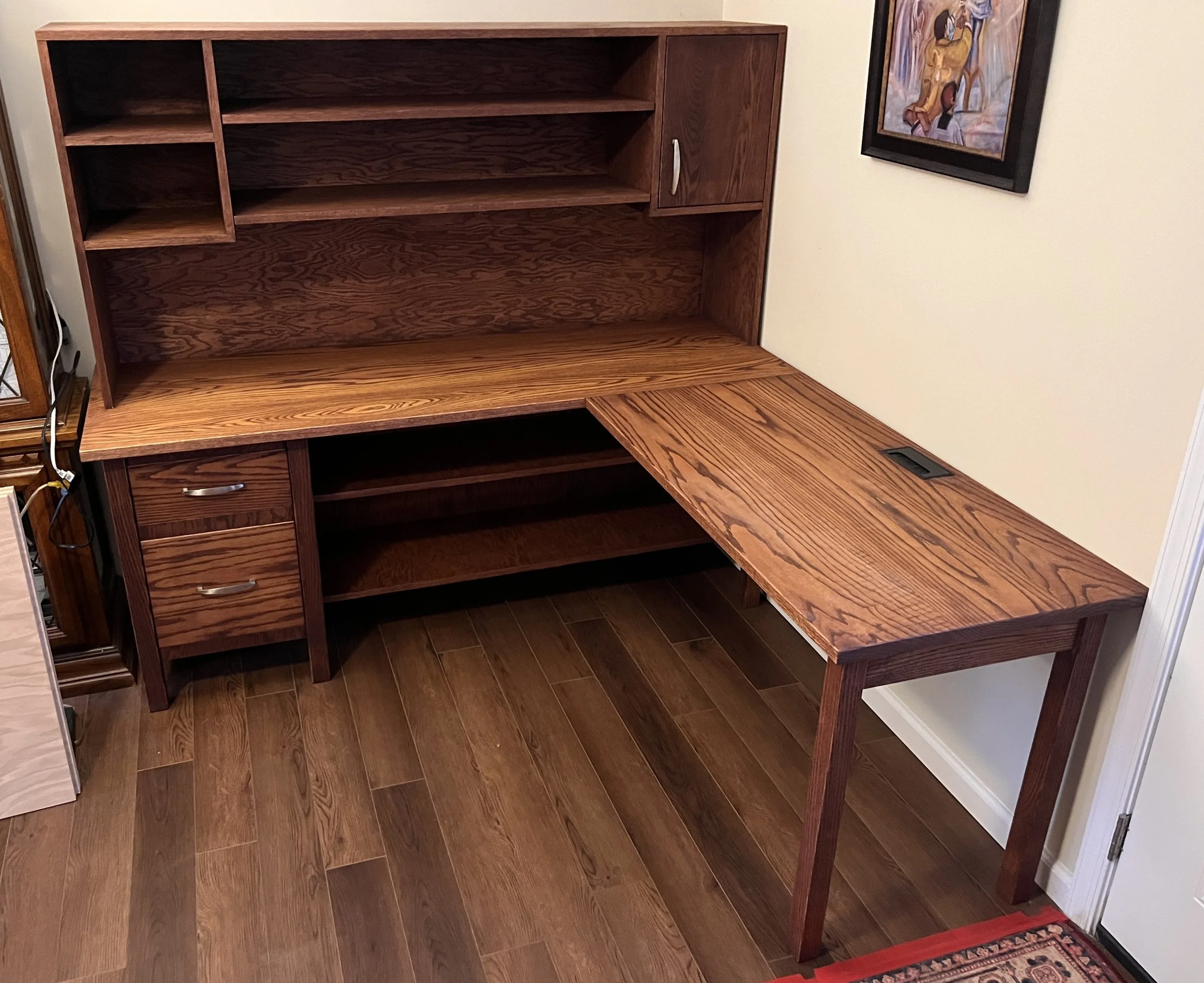 A wooden office desk with a hutch and a small cabinet on the right side, placed against a beige wall with a framed painting hanging nearby.
