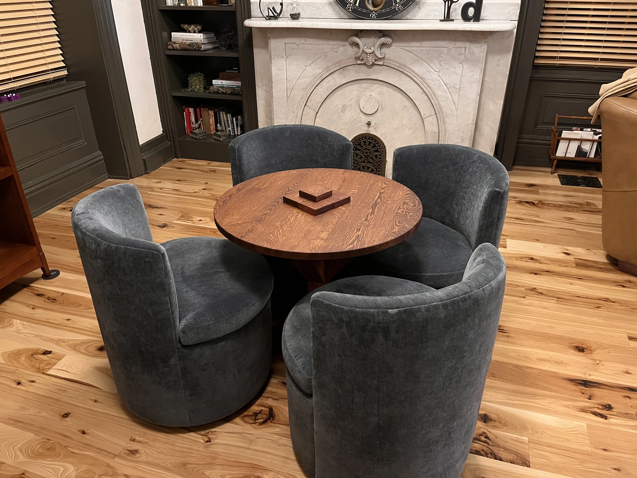 A round wooden table surrounded by four upholstered gray chairs in a living room with hardwood flooring, a fireplace, built-in bookshelves, and window blinds.