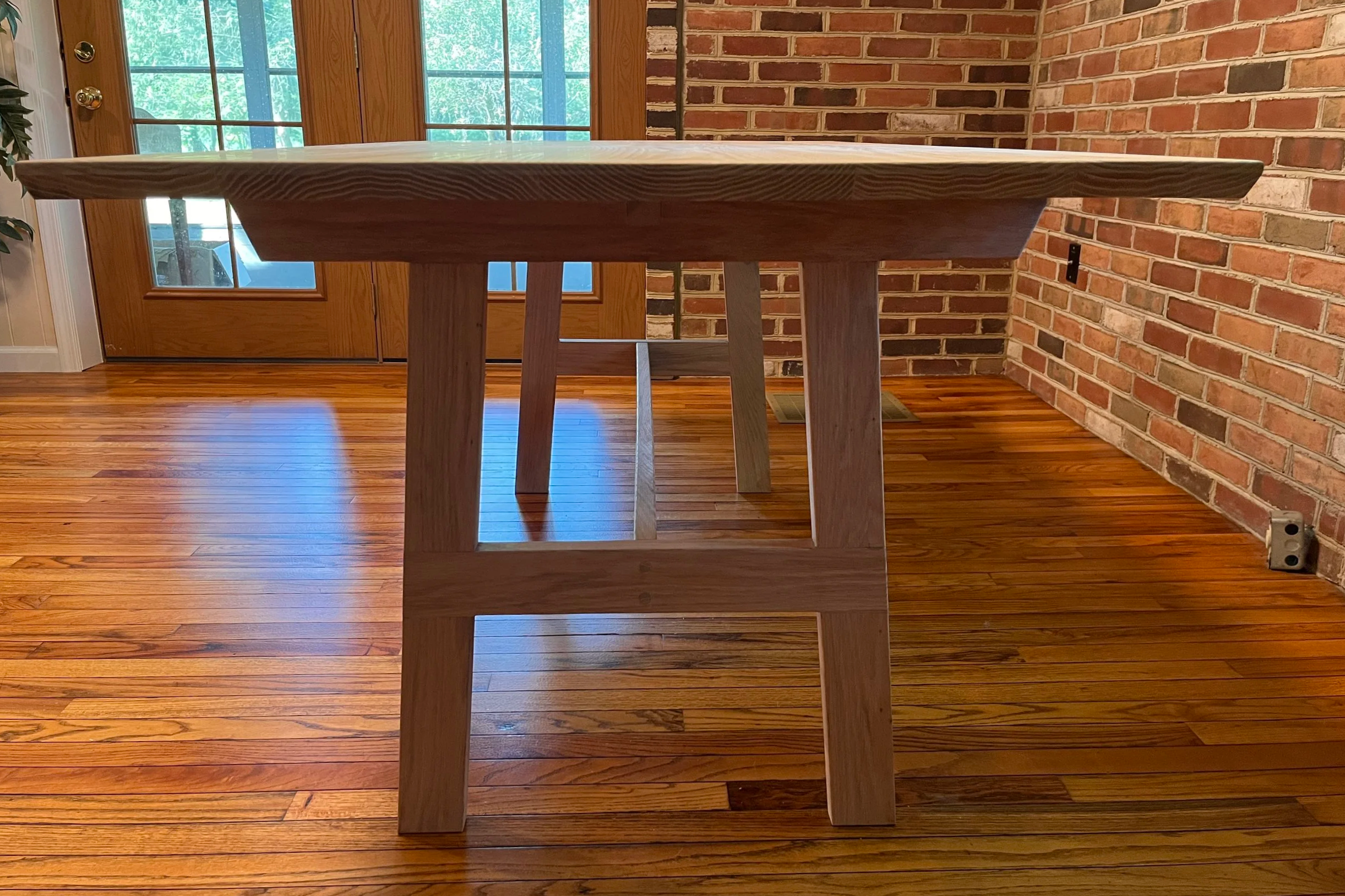 A wooden dining table with a natural wood finish, situated on a hardwood floor, against a brick wall and a glass door with wooden frames.