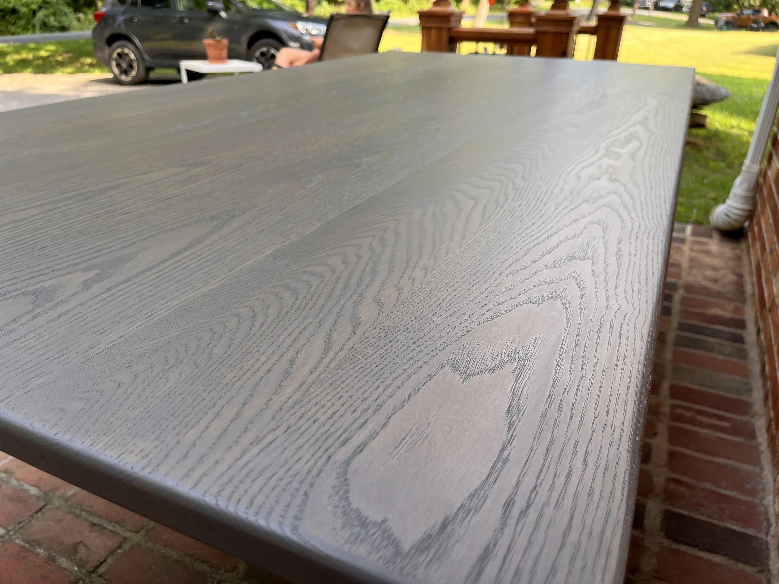 Close-up of a wooden outdoor table with a gray finish, showing wood grain pattern, situated on a brick patio.