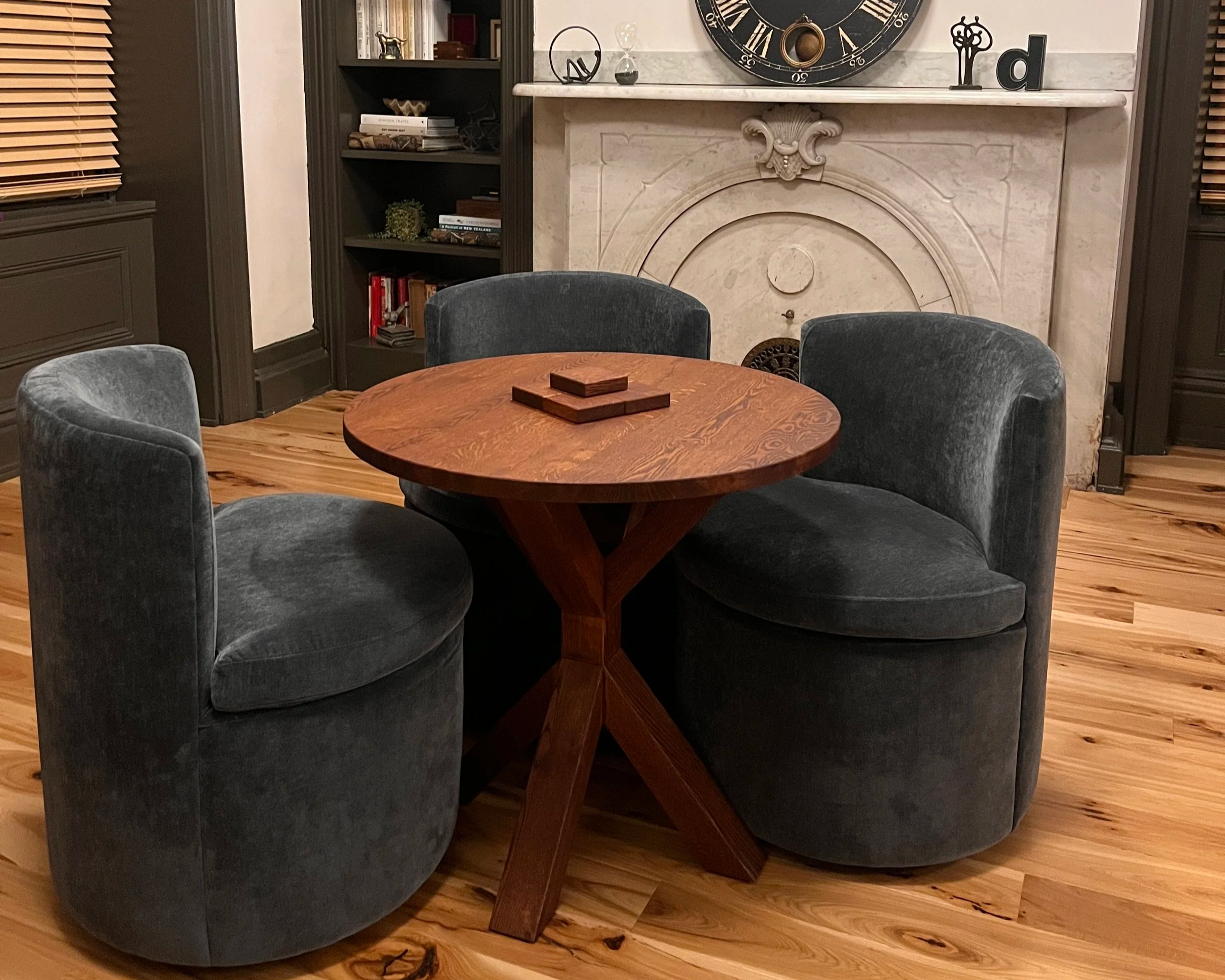 A round wooden table surrounded by four dark gray velvet chairs in a cozy room with wooden flooring, a fireplace with decorative molding, a black bookshelf filled with books, and a large black clock above the fireplace mantel.