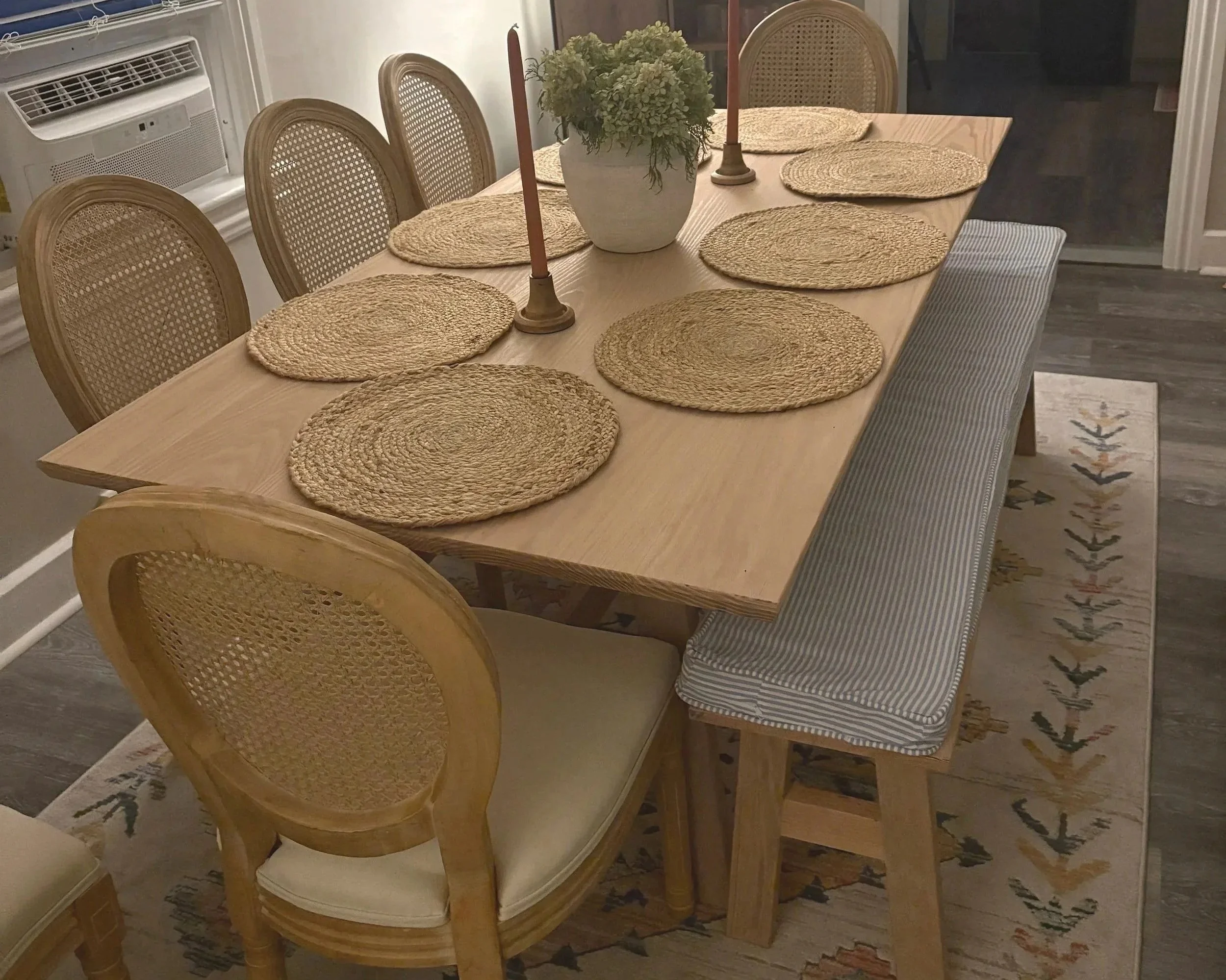 A wooden dining table set with eight woven placemats, two pink candles, and a potted plant centerpiece, surrounded by six chairs and a striped bench, on a patterned rug in a dining room.