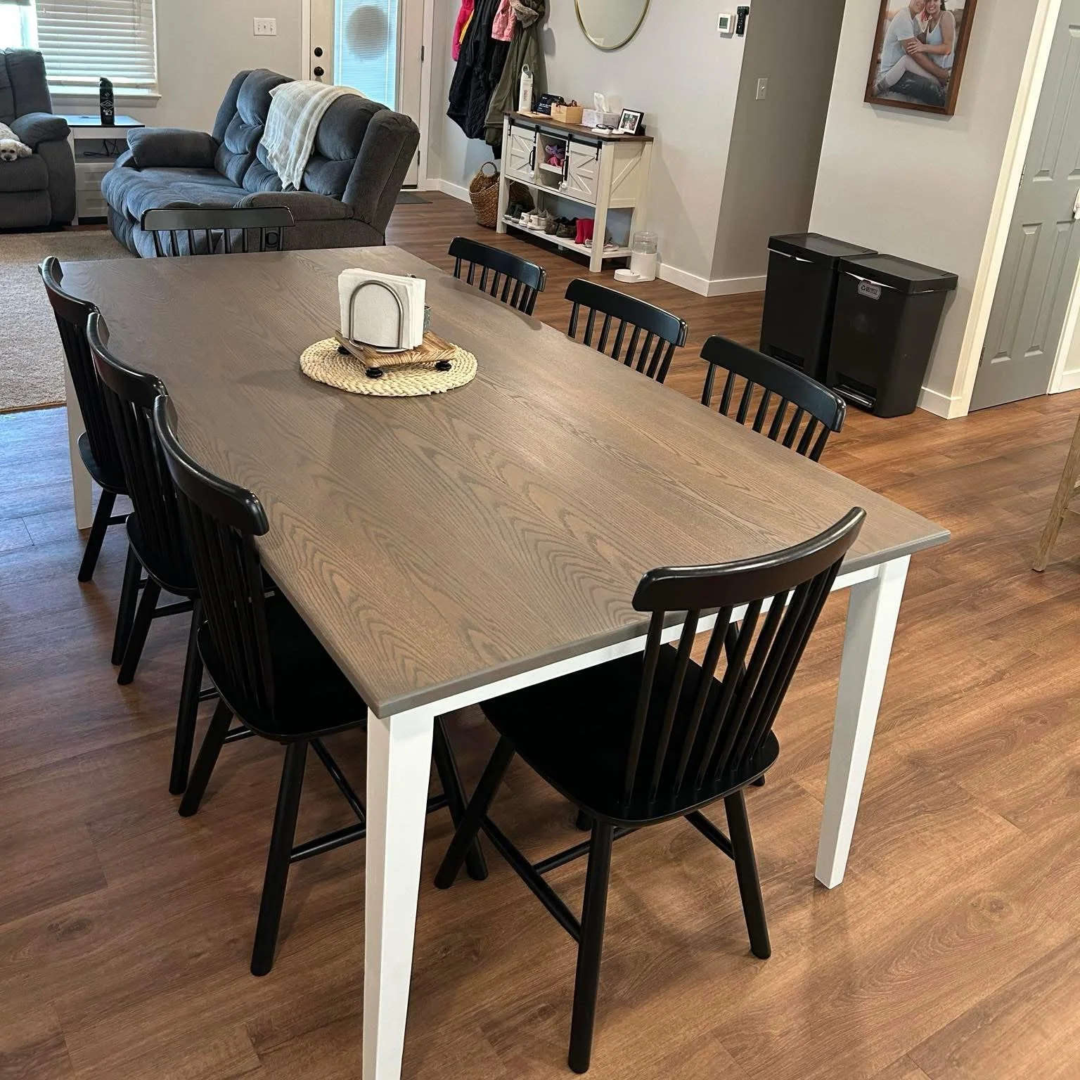 A dining table with black chairs in a living room with two gray couches and a chandelier overhead.