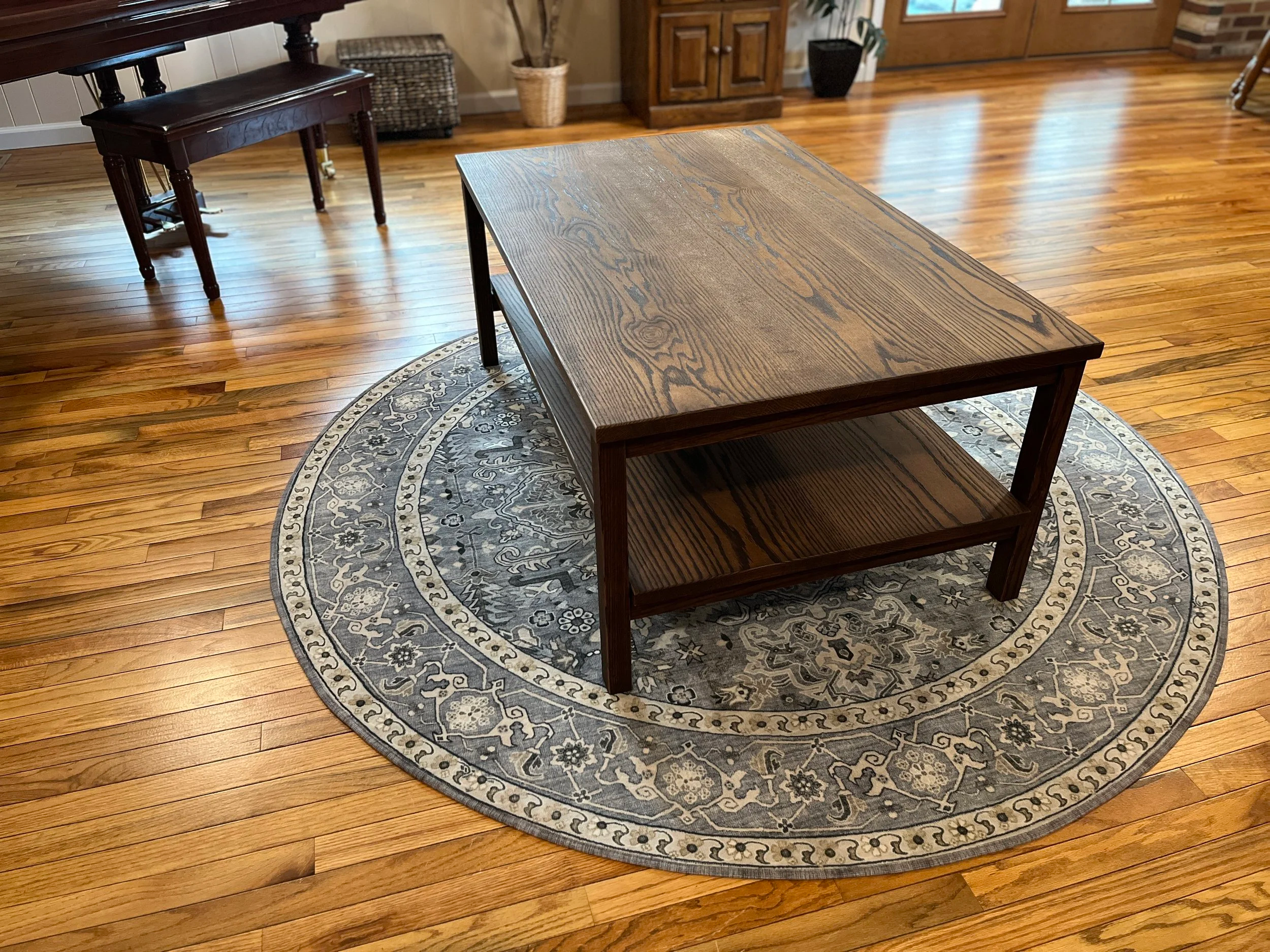 A wooden coffee table with a lower shelf on a round patterned rug on hardwood floor in a living room.