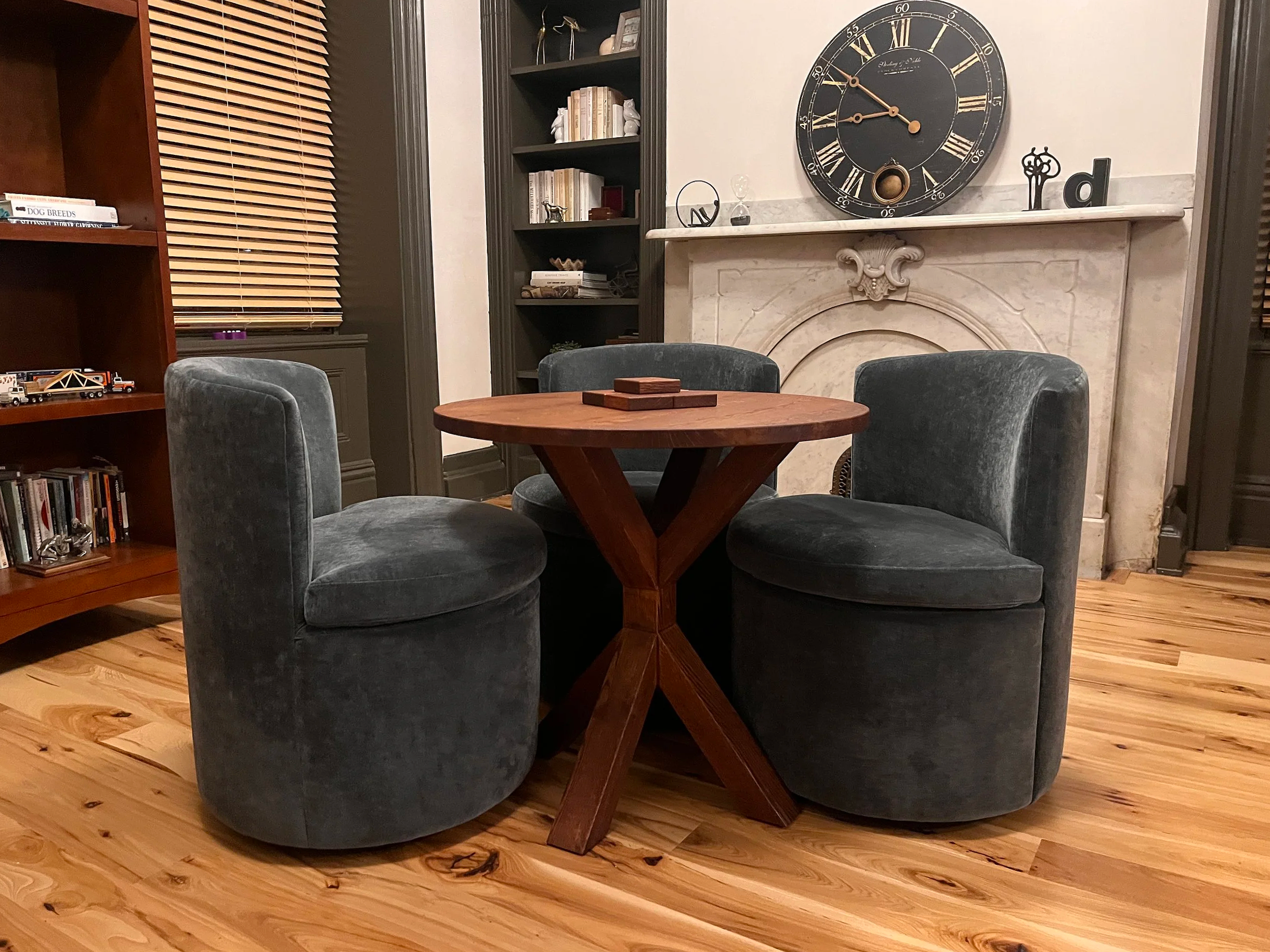 A cozy dining room with a round wooden table surrounded by four velvet upholstered chairs. Behind the table is a fireplace with a decorative mantel and a large wall clock above it. To the left is a bookshelf filled with books and decorative items, and a closed window with wooden blinds.