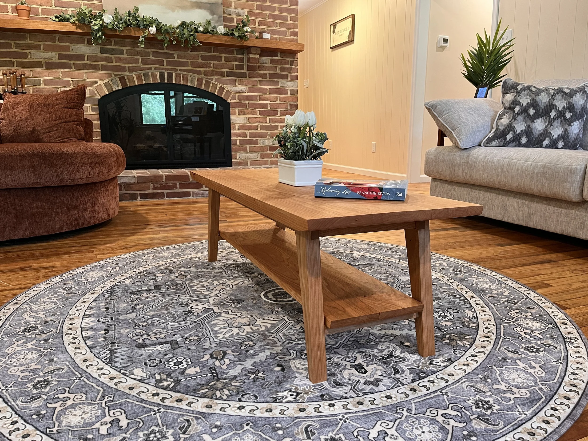 Living room with brick fireplace, wooden coffee table, patterned rug, beige and gray couches, houseplant, and decorative items.