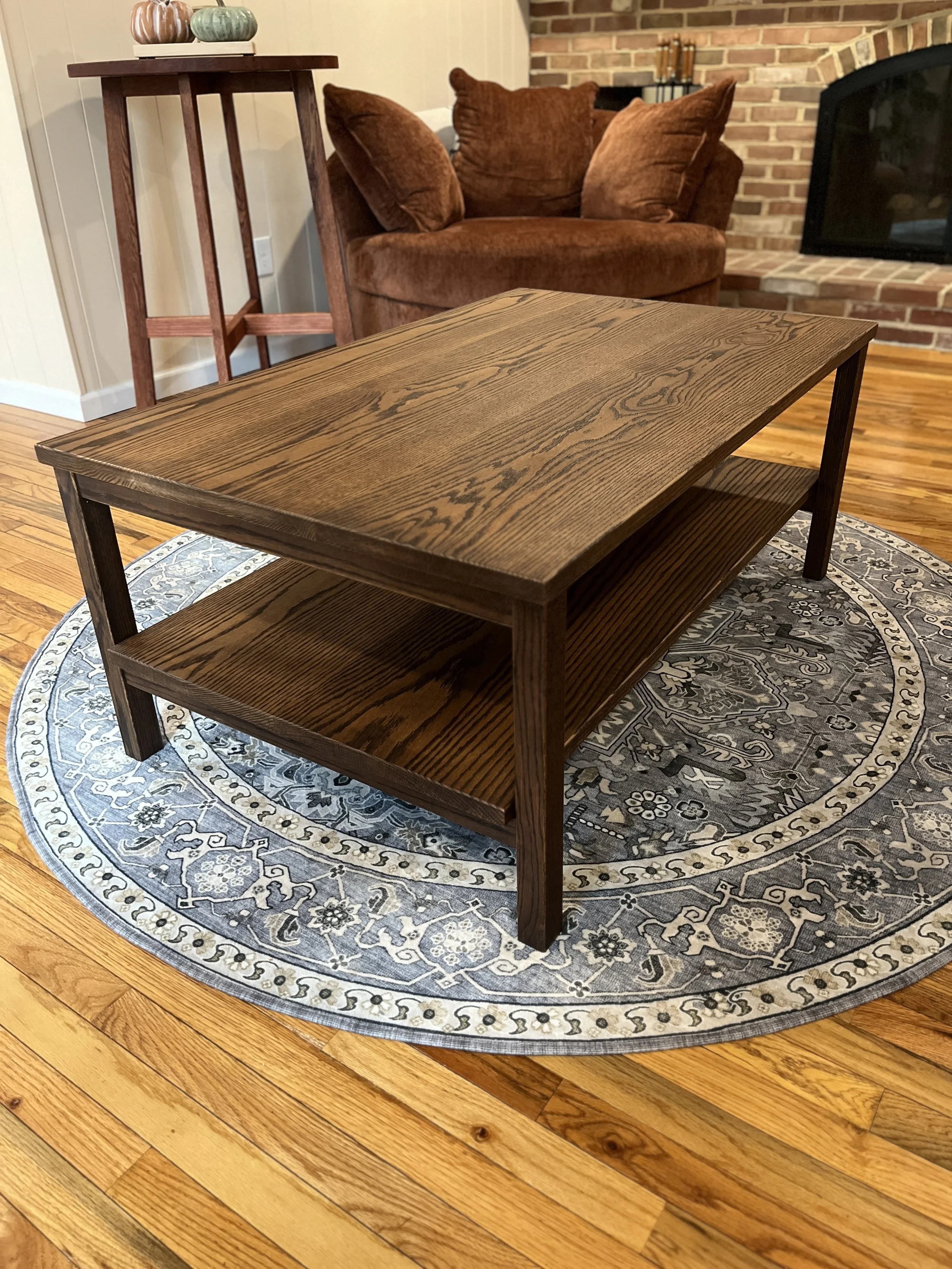 A wooden coffee table on a round decorative rug in a living room with hardwood floors, a brown armchair, and a brick fireplace.