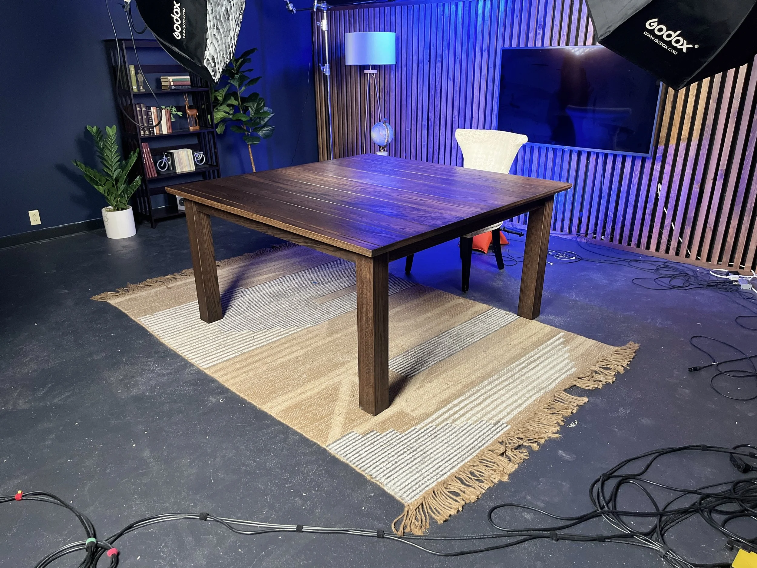 A wooden conference table with a white cushioned chair behind it, placed on a patterned rug in a room with dark walls, a bookshelf, potted plants, a standing lamp, and a wall-mounted television. Photography equipment and cables are visible on the flo