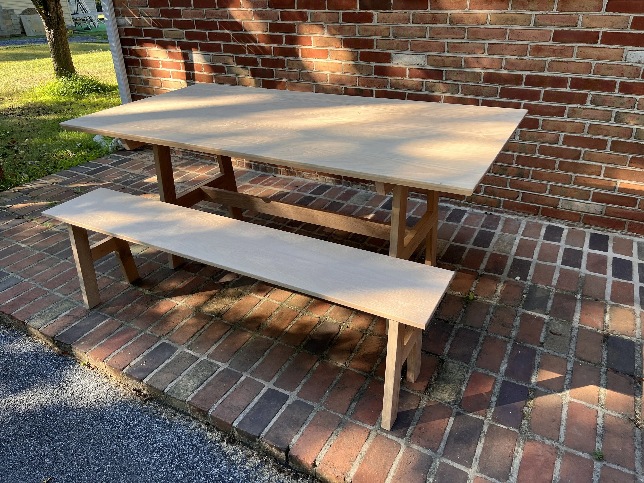 Wooden table with a matching bench on a brick patio next to a brick wall, with sunlight and shadows.