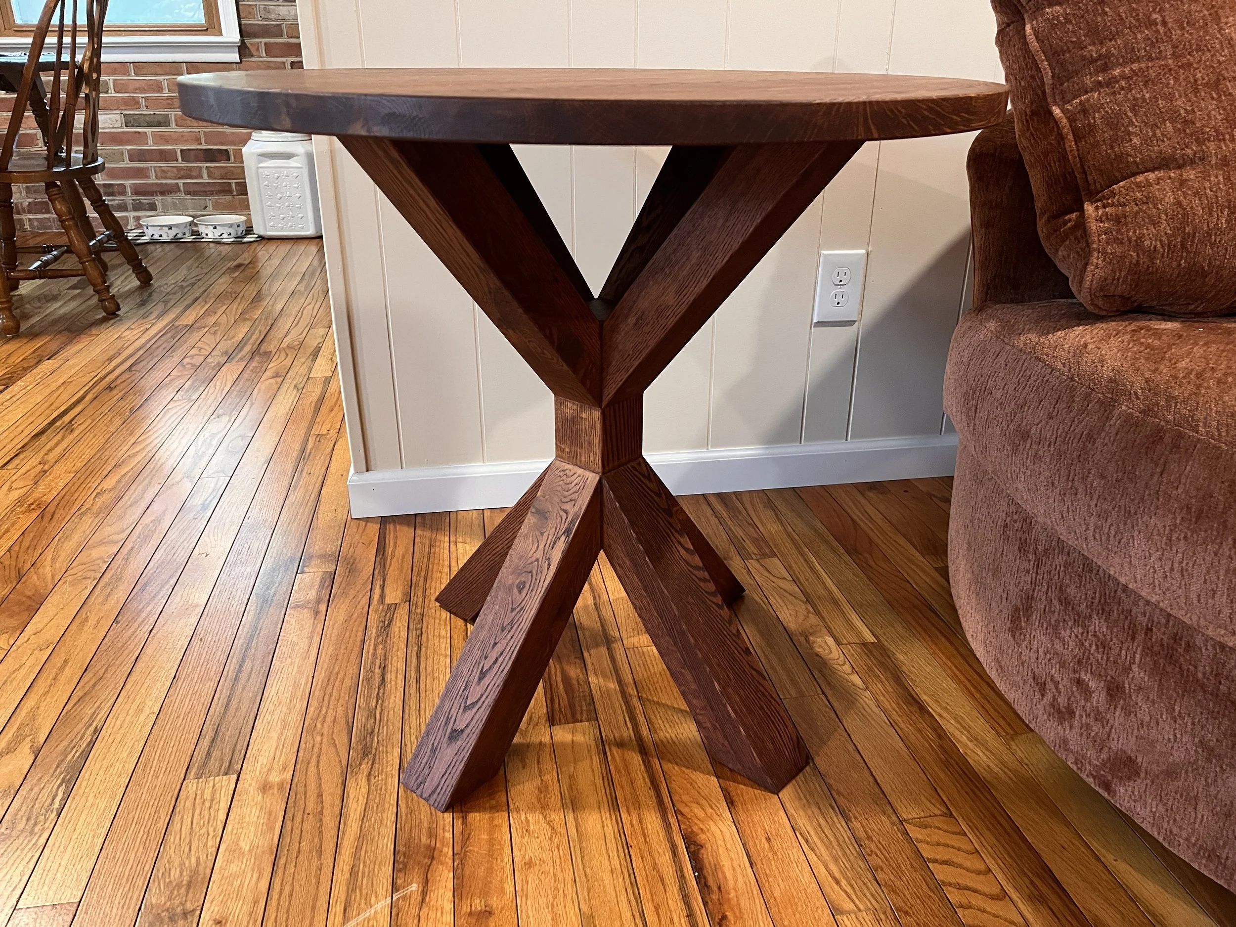 A wooden round table with a cross-legged base in a room with hardwood floors and a brown upholstered sofa.