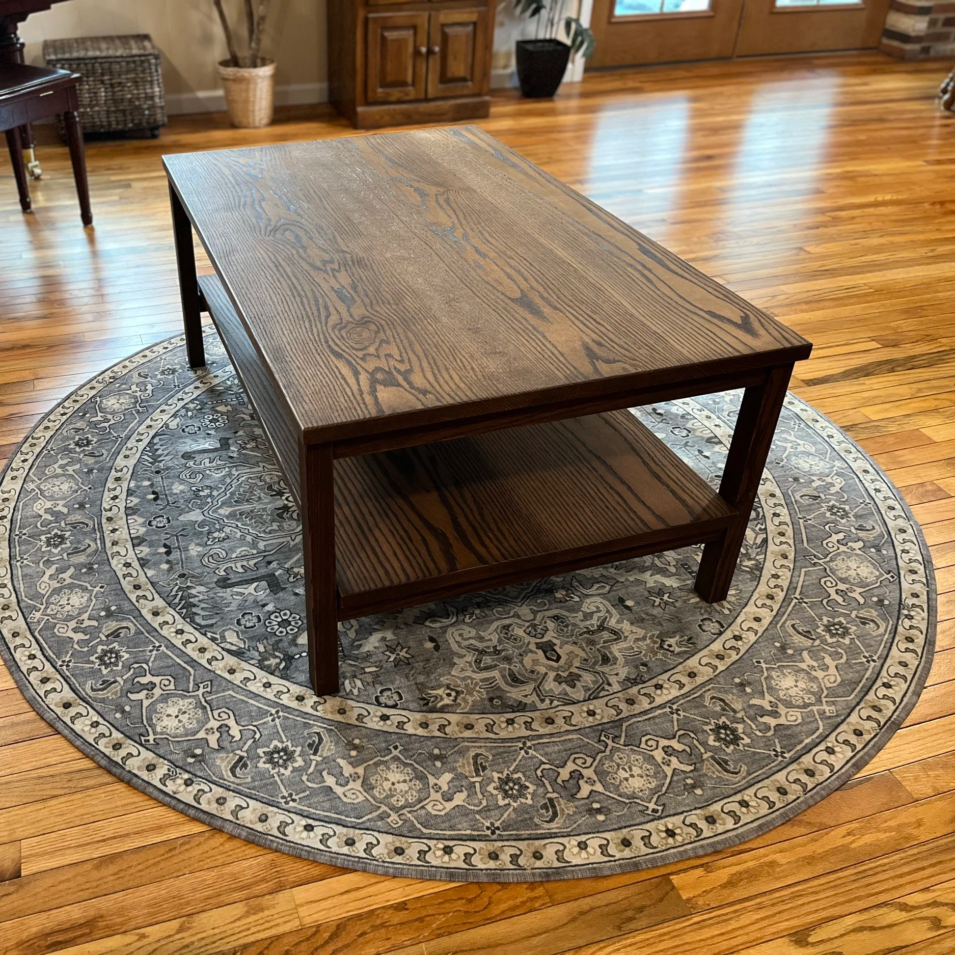 A wooden coffee table with two shelves placed on a decorative round area rug with a floral and geometric pattern, set on a hardwood floor in a living room. In the background, there are plants, a wooden cabinet, and a door.