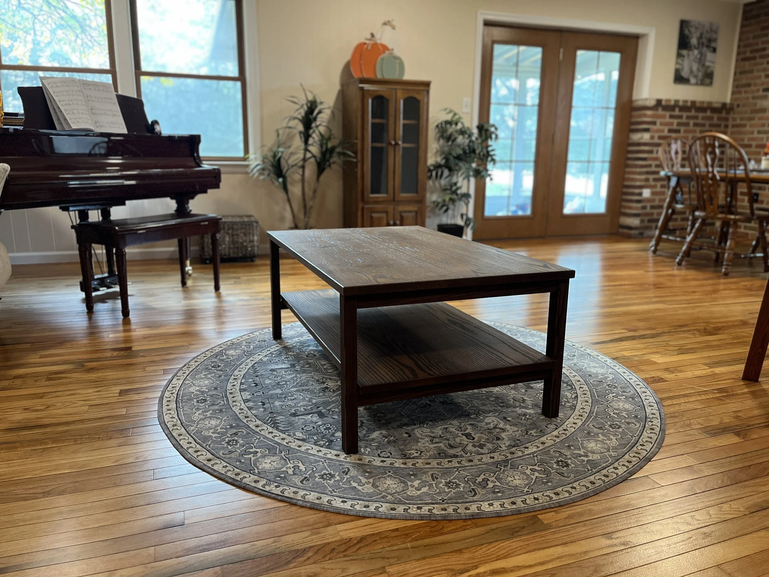 Living room with wooden flooring, a round patterned rug, a wooden coffee table, a piano with sheet music, plants, a tall cabinet, and a bar-height dining table with chairs near a glass door.