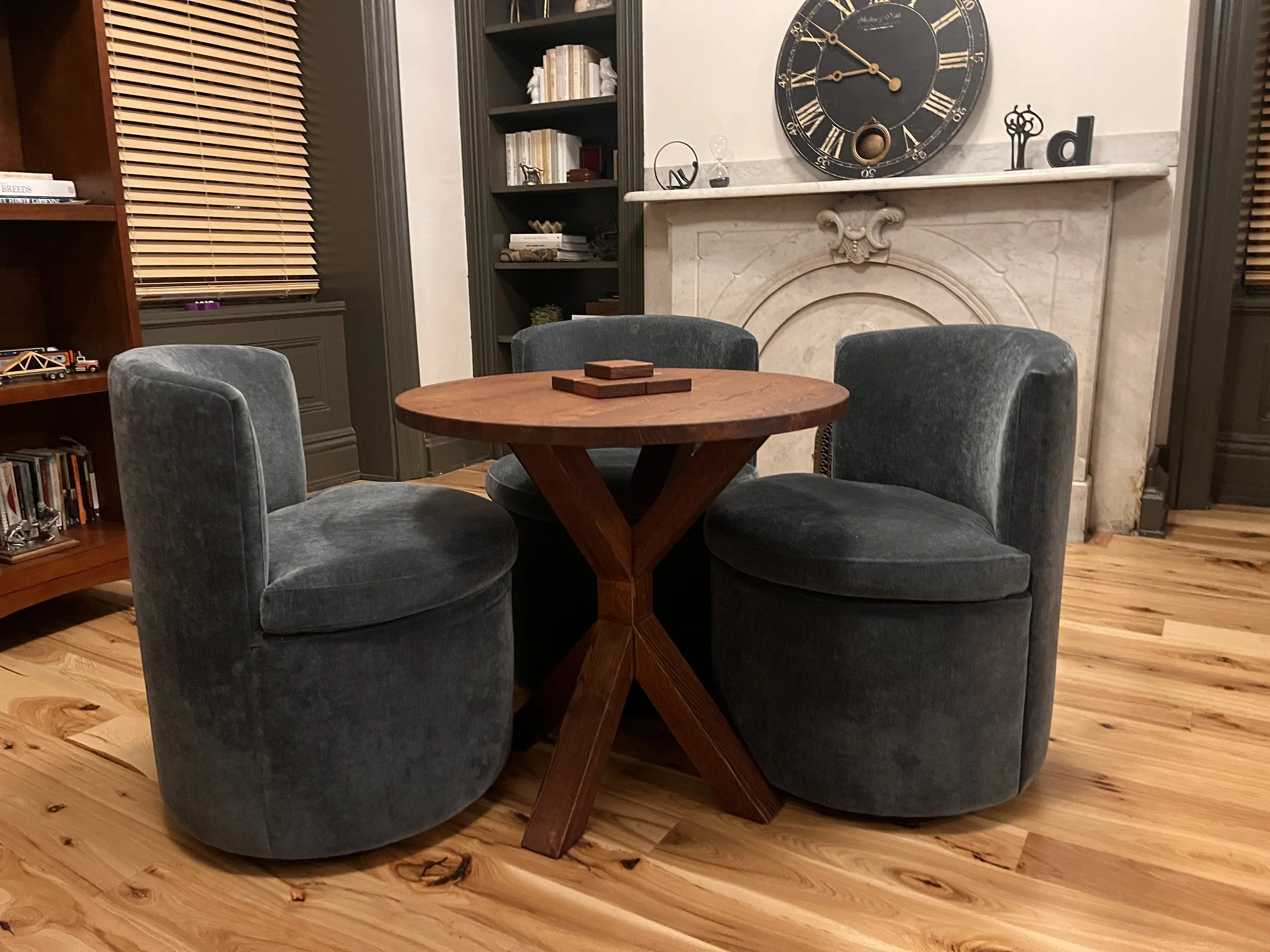 A cozy living room corner with a round wooden table surrounded by three upholstered gray chairs, a fireplace with a decorative mantle, a large wall clock, bookshelves filled with books, and wooden floors.