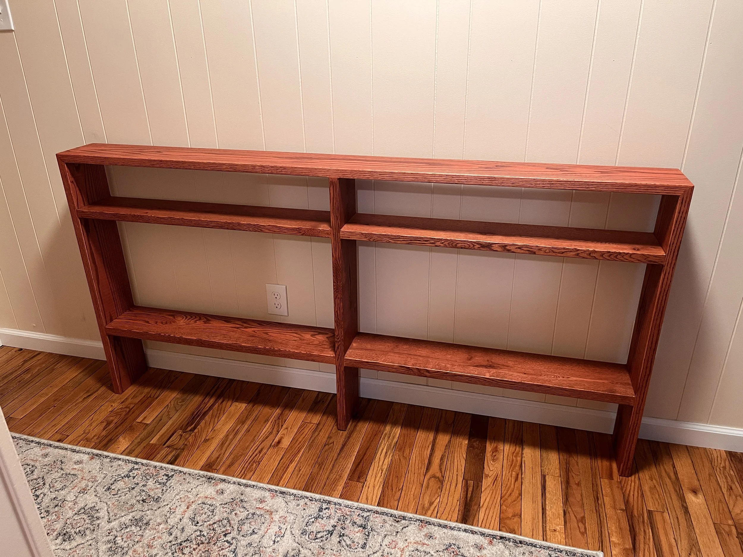 Empty wooden shelf unit with three levels and vertical divider, placed against a wall with wooden paneling, on a hardwood floor near a rug.