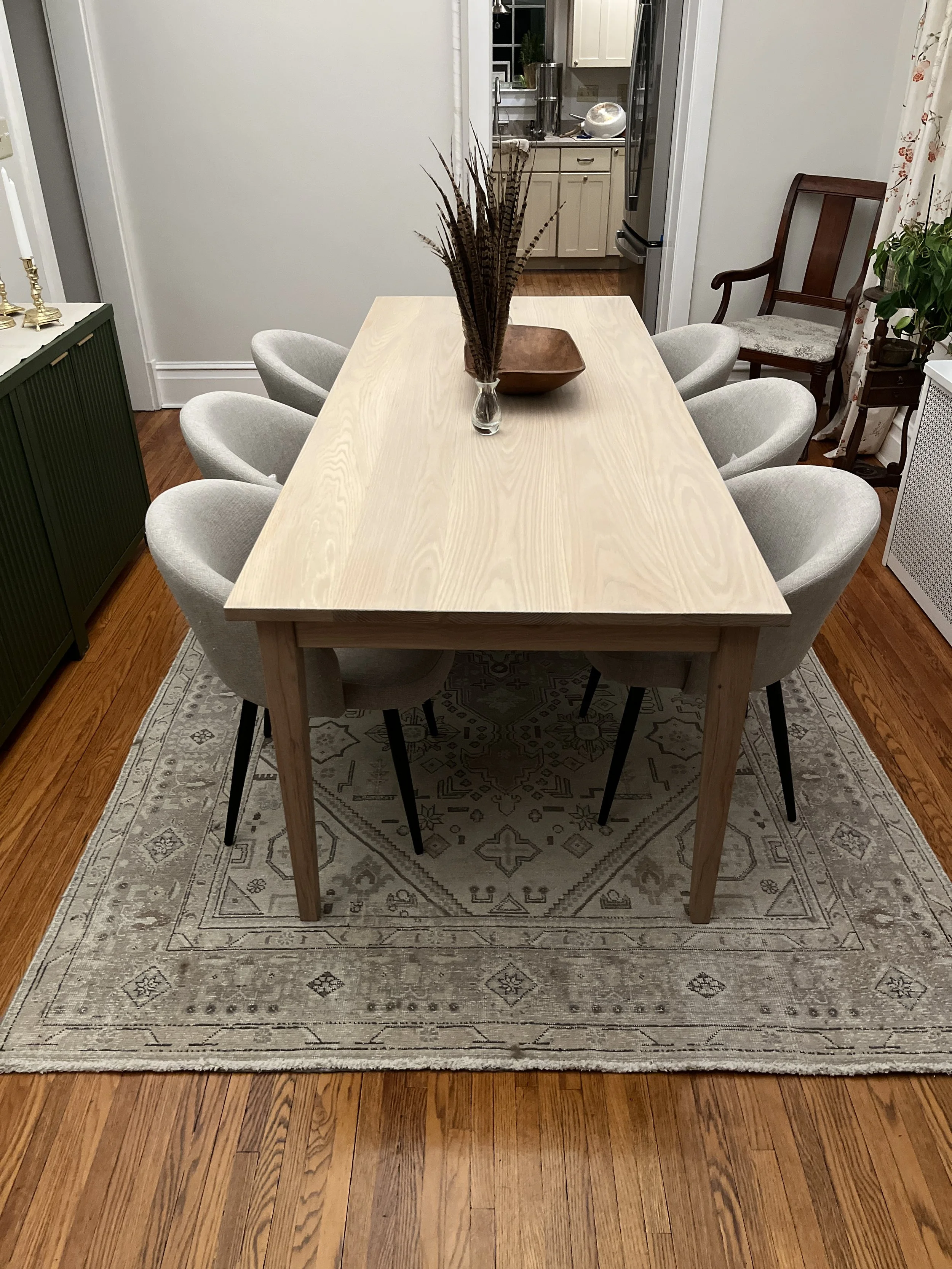 A dining room with a wooden table, six light-colored upholstered chairs, a beige rug, a vase with dried plants, and a wooden bowl on the table. There is a green sideboard on the left and a white curtain with floral pattern on the right. The backgroun