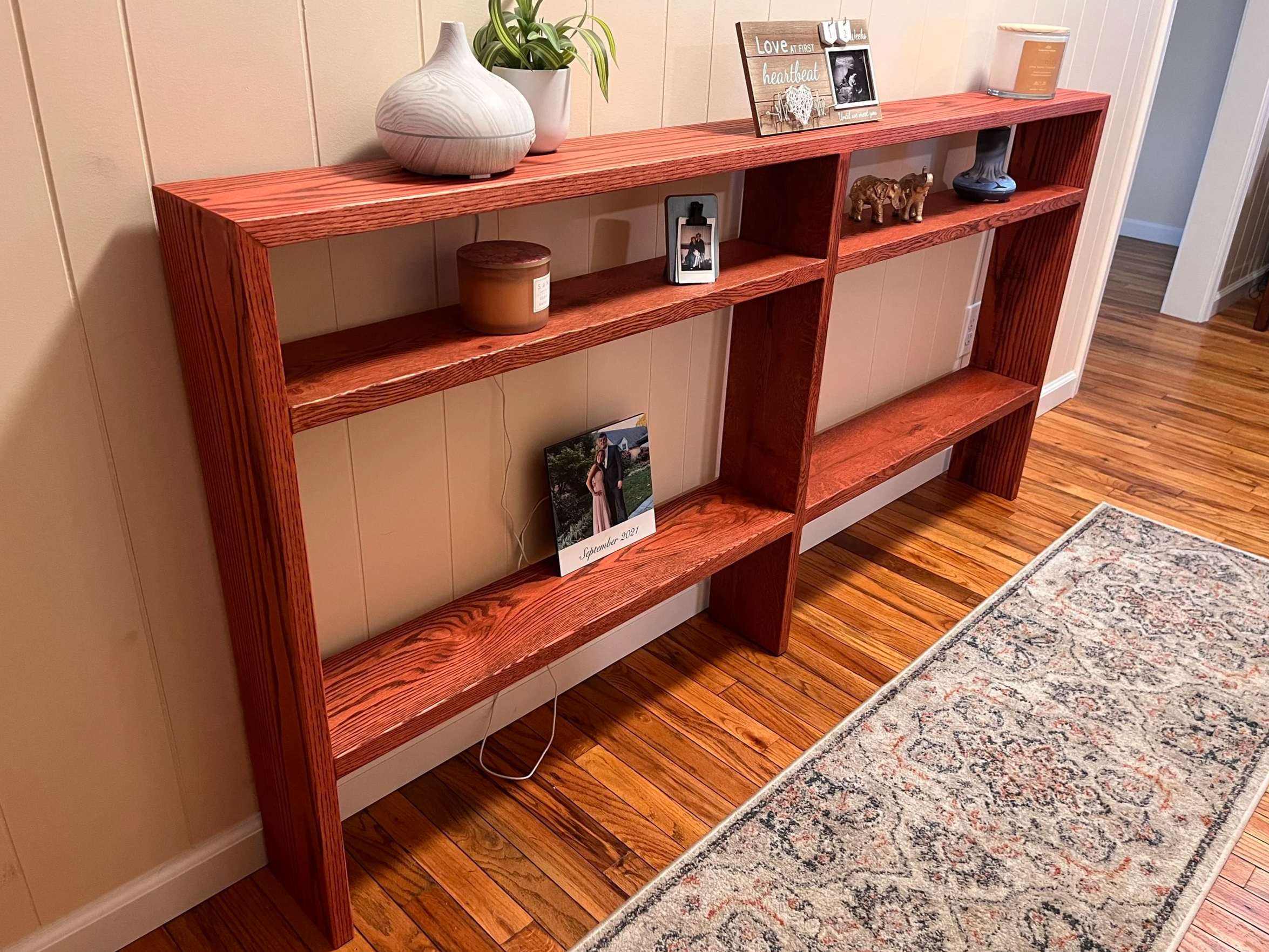 A wooden bookshelf with multiple shelves against a beige wall, decorated with a white vase with green plants, a candle, framed photos, small figurines, and a decorative plate, with a patterned rug on a hardwood floor in front.