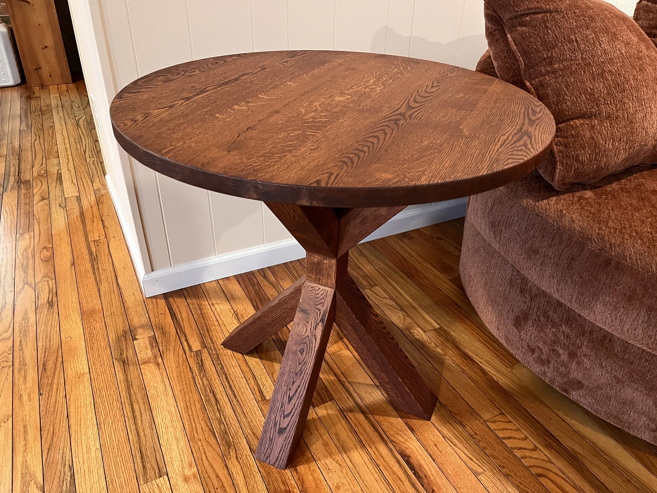 A round wooden side table next to a red upholstered sofa on a hardwood floor.