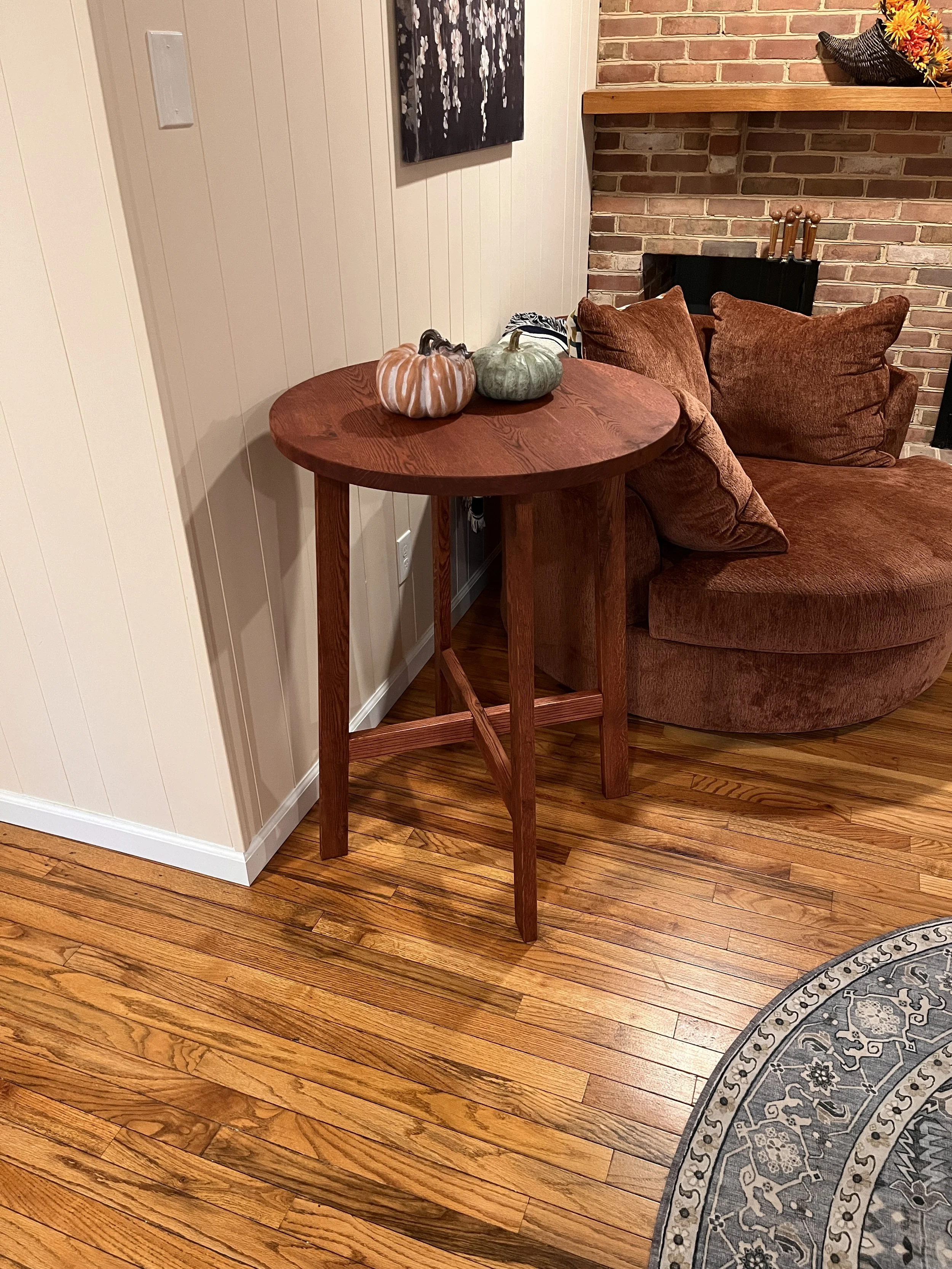 A round wooden side table with decorative pumpkins on top, situated next to a sectional sofa with brown cushions in a living room with hardwood floors and a brick fireplace.