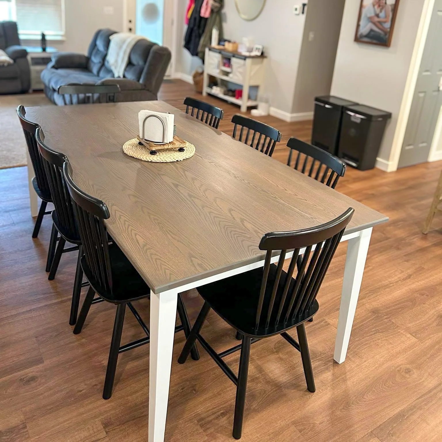 A wooden dining table surrounded by six black chairs in a room with hardwood floors, with a living area in the background featuring a sofa, a side table, and a small white shelf.