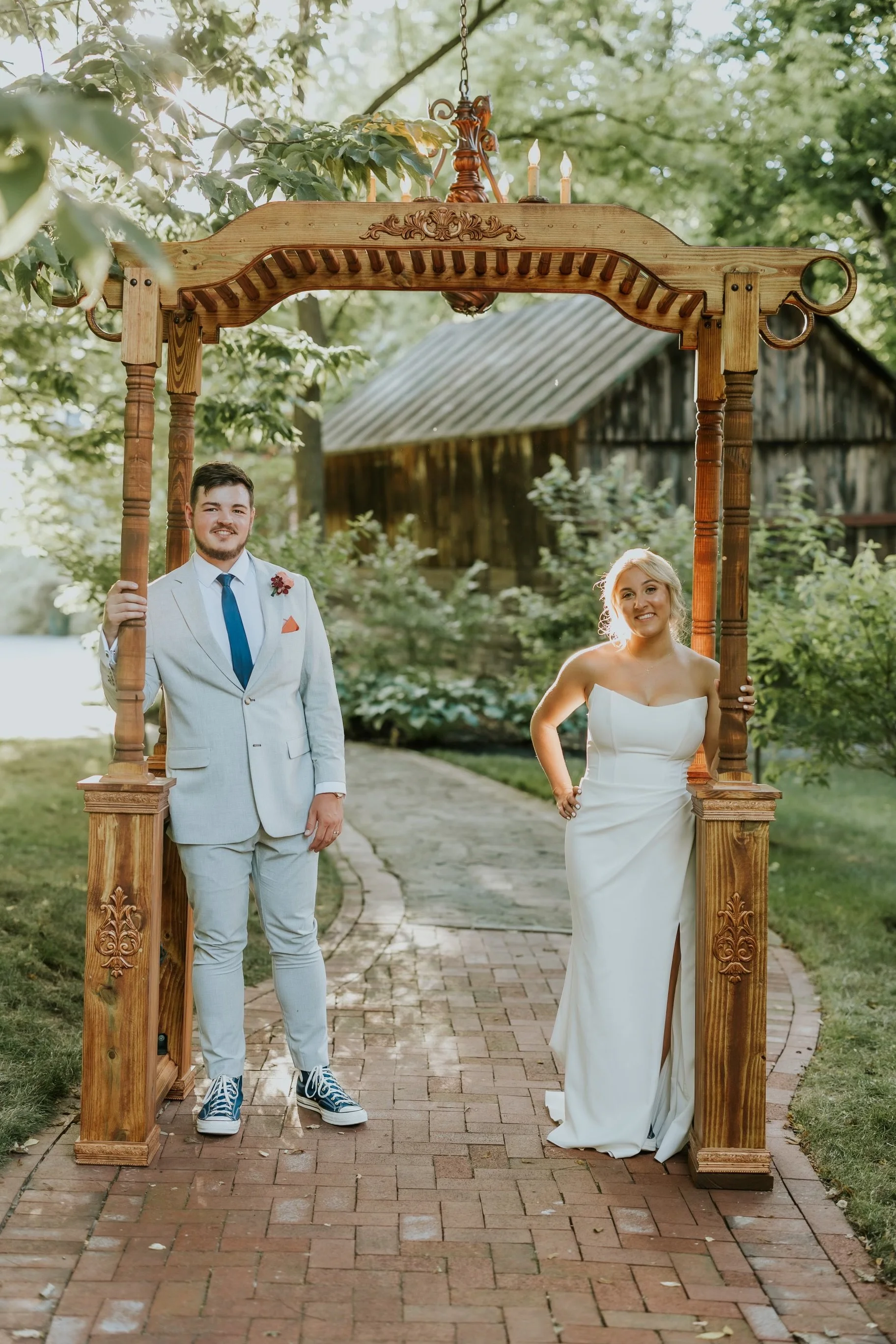 A bride and groom standing under a wooden archway outdoors during their wedding, with sunlight filtering through trees. The groom wears a light gray suit with a blue tie, and the bride wears a white strapless gown.