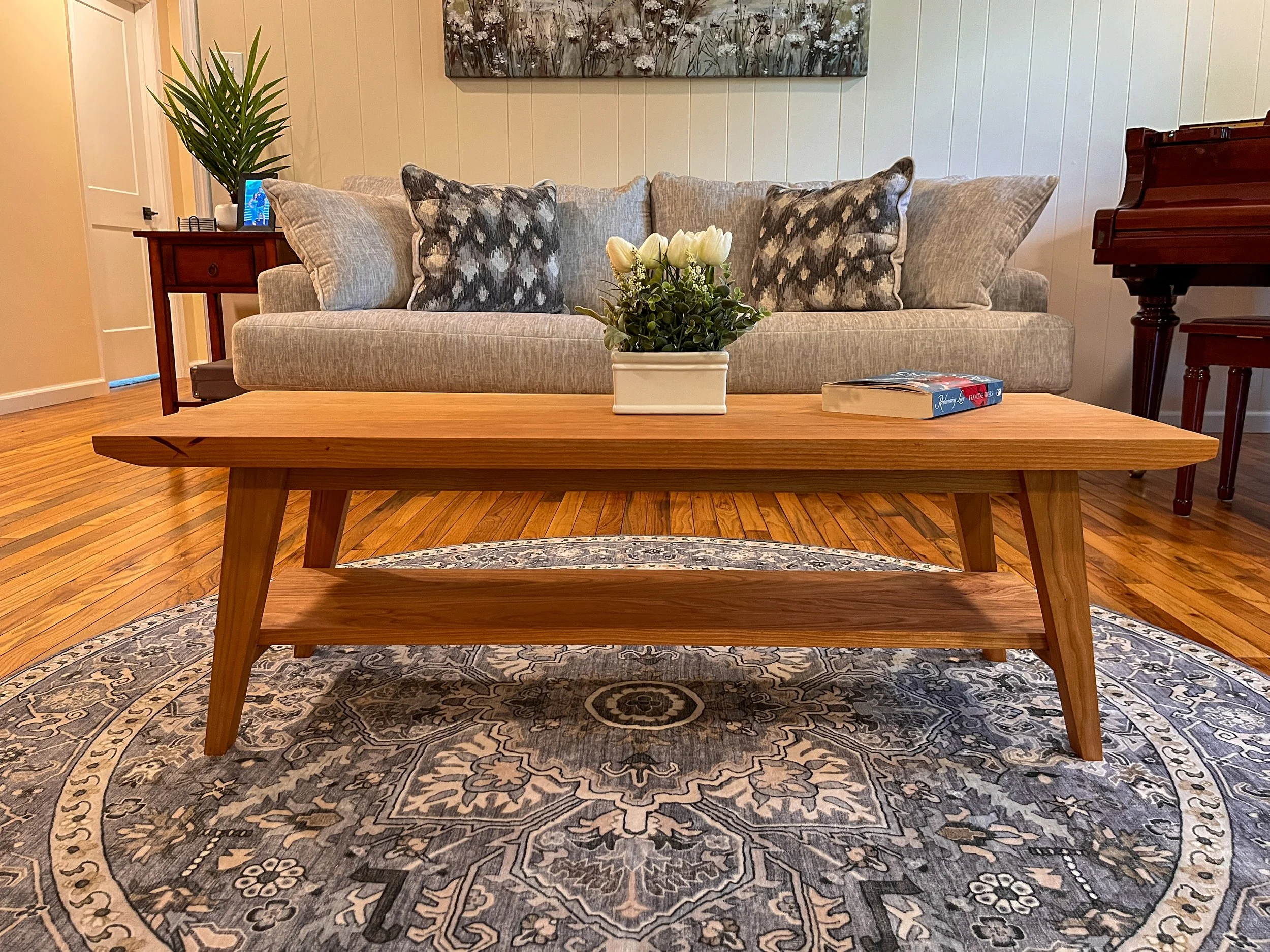 Living room with a wooden coffee table, a beige sofa with patterned pillows, a potted plant, a book, and a grand piano in the background, with a round rug on a wooden floor.