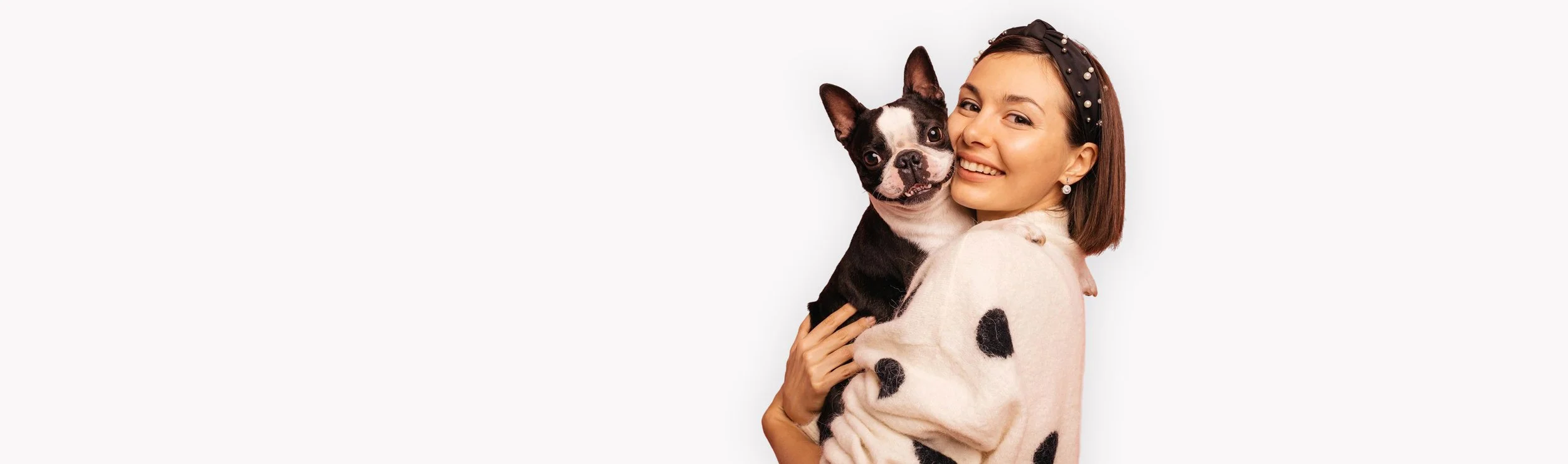 A woman holding a black and white French Bulldog, both smiling, woman wearing a beige sweater with black spots and a black headband with pearls.