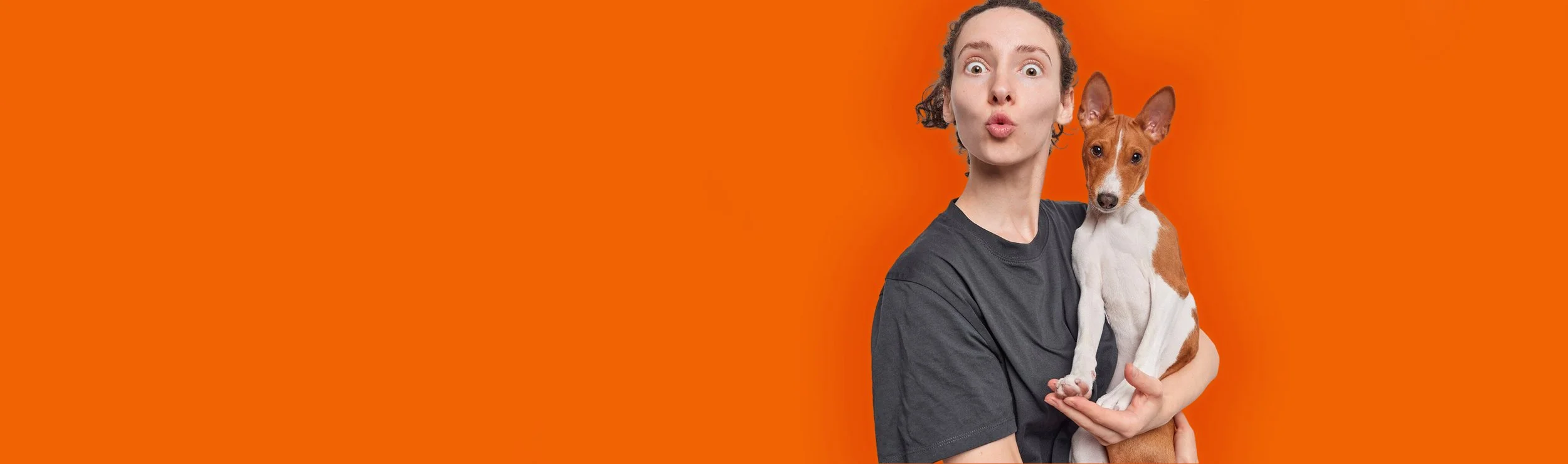 A woman with curly hair making a surprised expression while holding a small brown and white dog against an orange background.