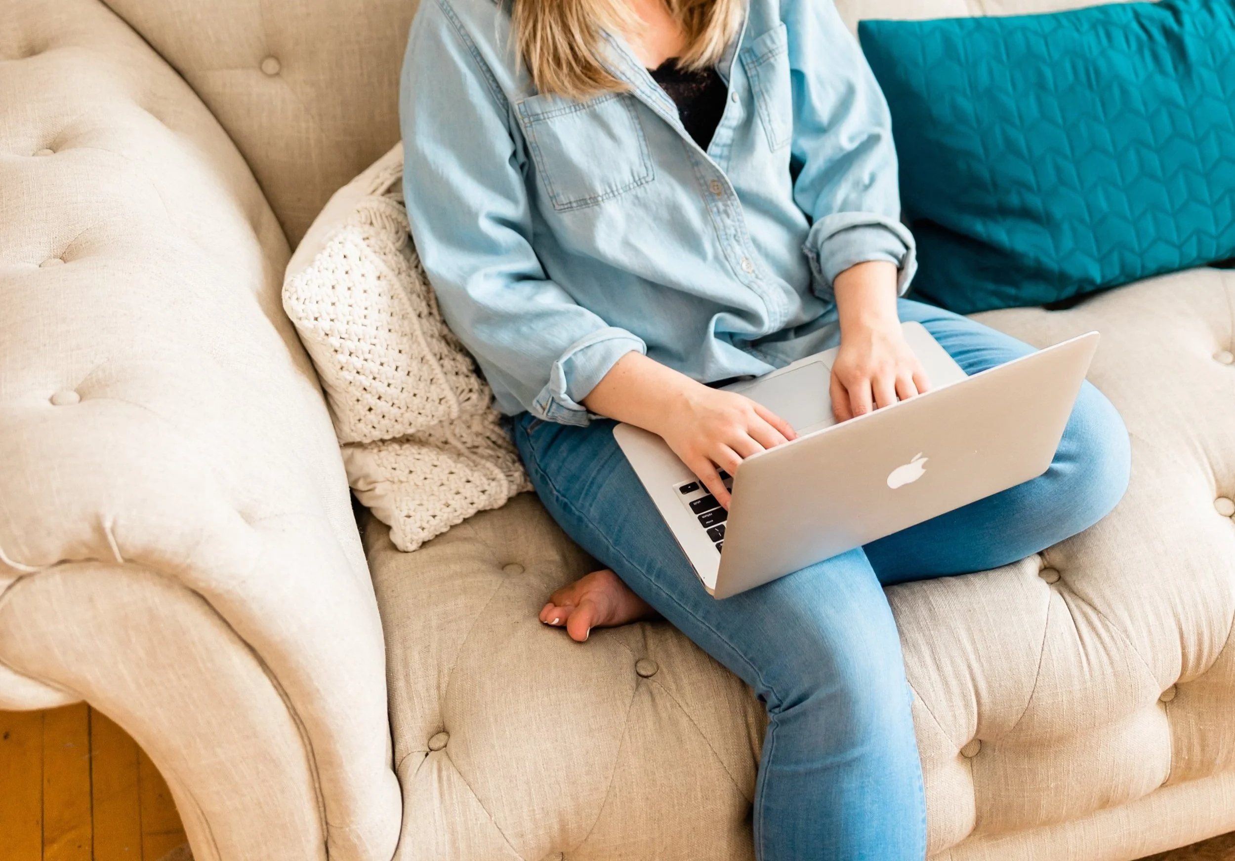 A person sitting cross-legged on a beige tufted sofa, using a silver MacBook laptop, with a white knitted pillow and a blue quilted pillow nearby.
