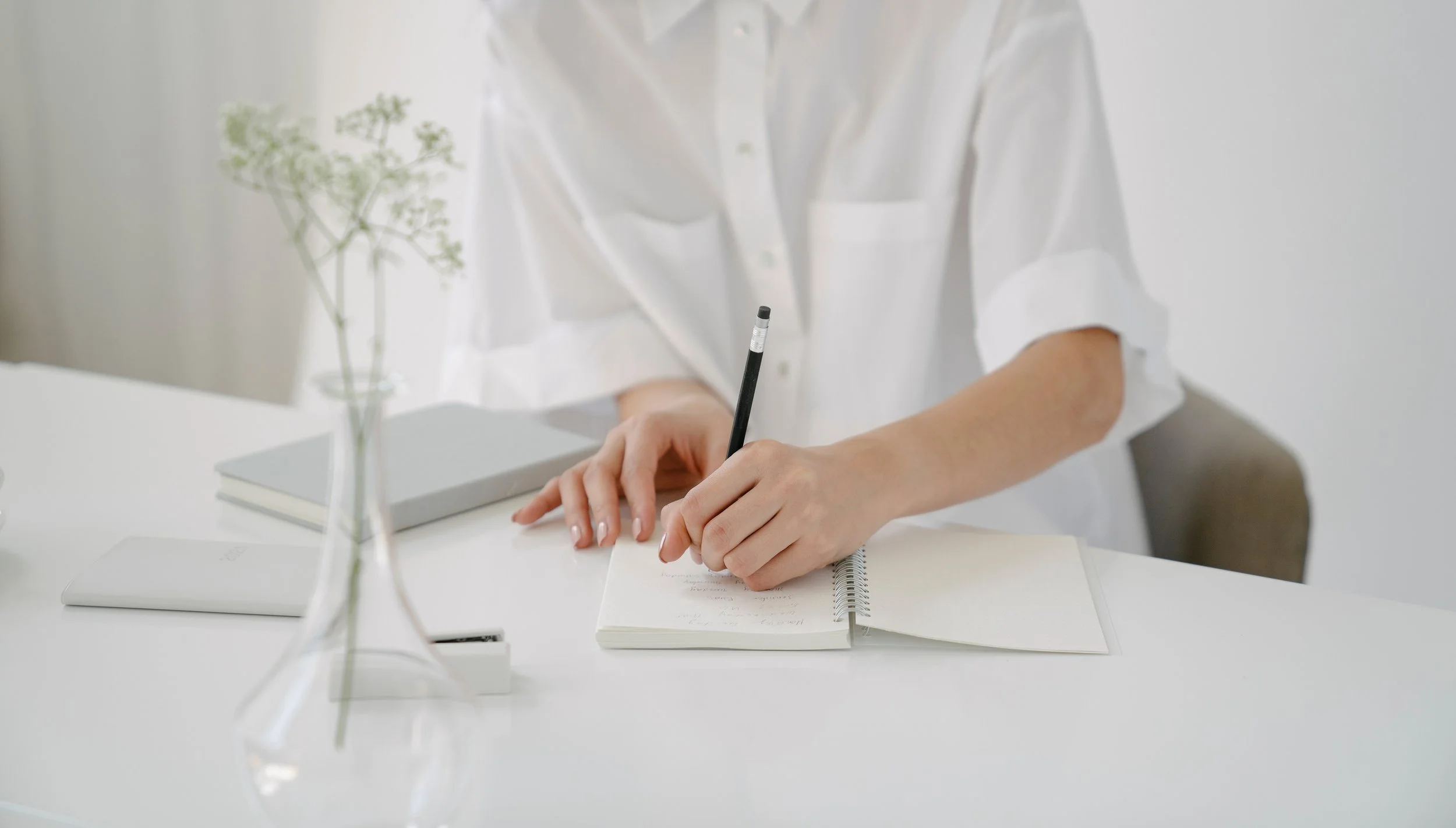 Person in white shirt writing in a notebook at a white desk with a laptop, closed notebook, and a glass vase with a plant.