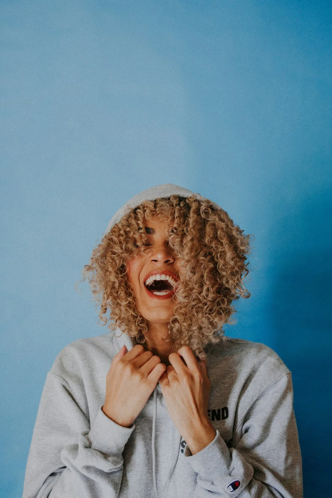 A woman with curly blonde hair, wearing a gray hoodie, smiling and laughing against a blue background.