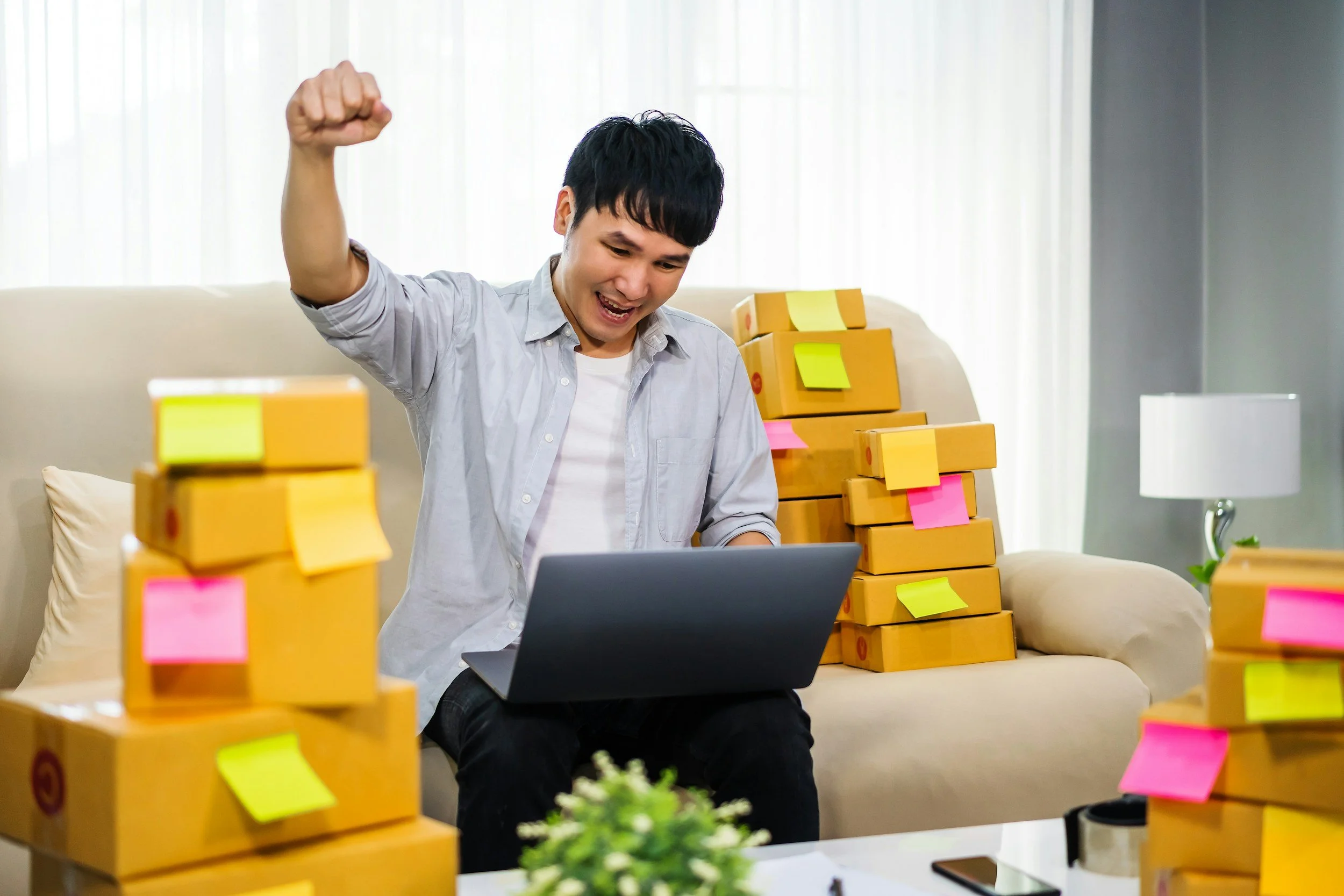 A young man sitting on a beige sofa with a laptop in his lap, surrounded by stacks of yellow boxes with colorful sticky notes, celebrating with a raised fist, in a bright living room.