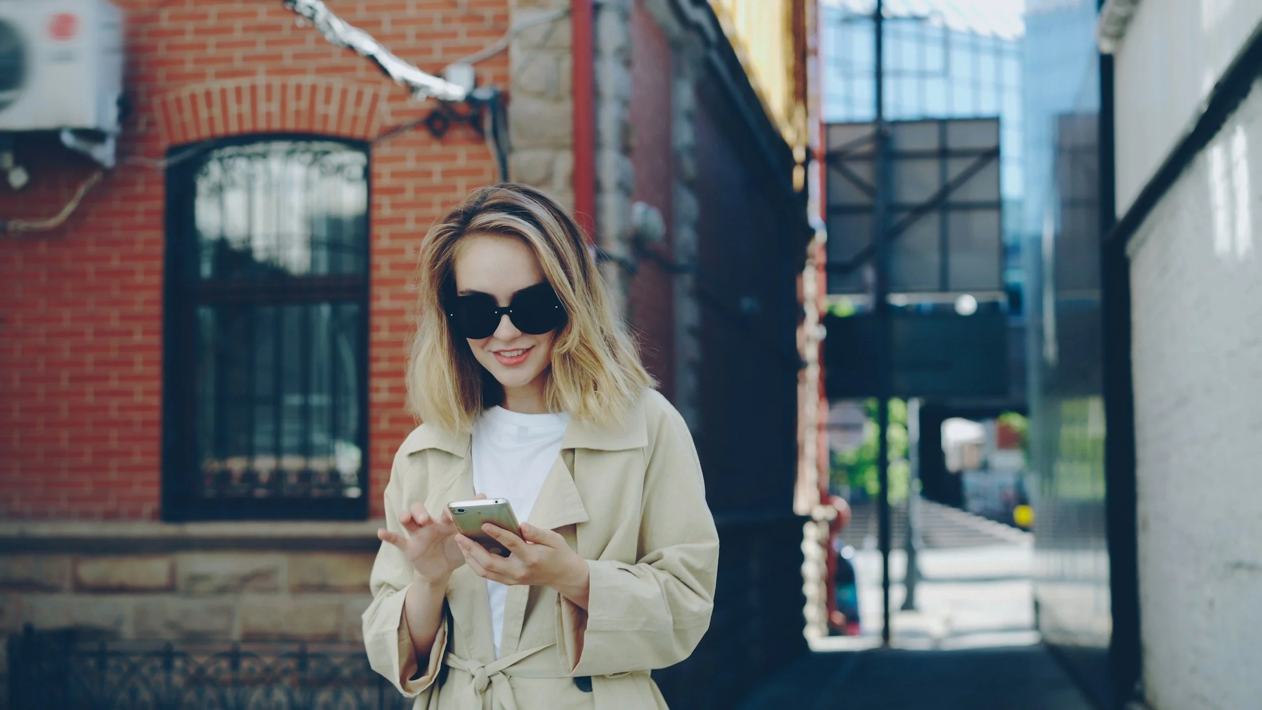 A woman with blonde hair wearing sunglasses and a beige coat, smiling and looking at her smartphone while standing in an alleyway with brick and light-colored buildings.