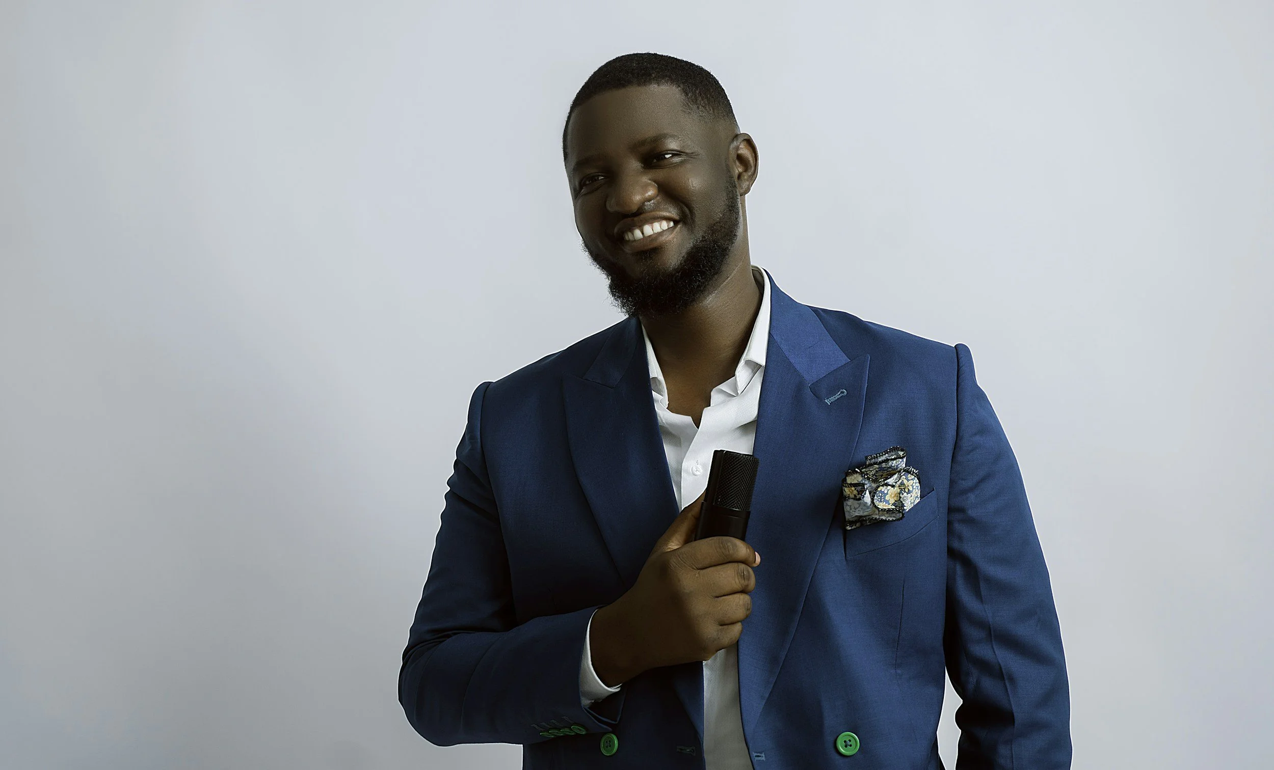 A man in a blue suit holding a microphone, smiling, against a plain white background.